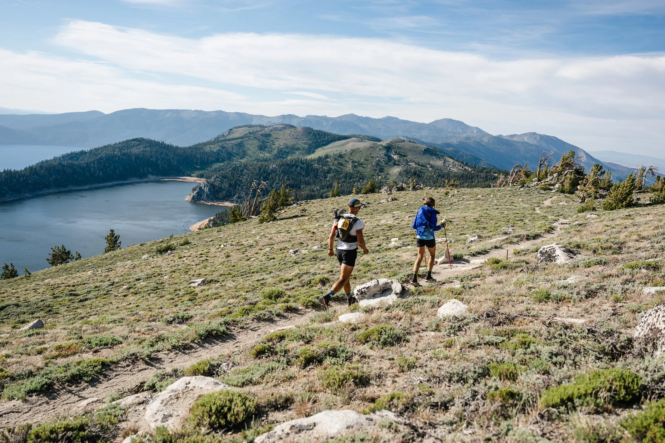 Two hikers walking on a trail through a hilly landscape with a lake and mountains in the distance.