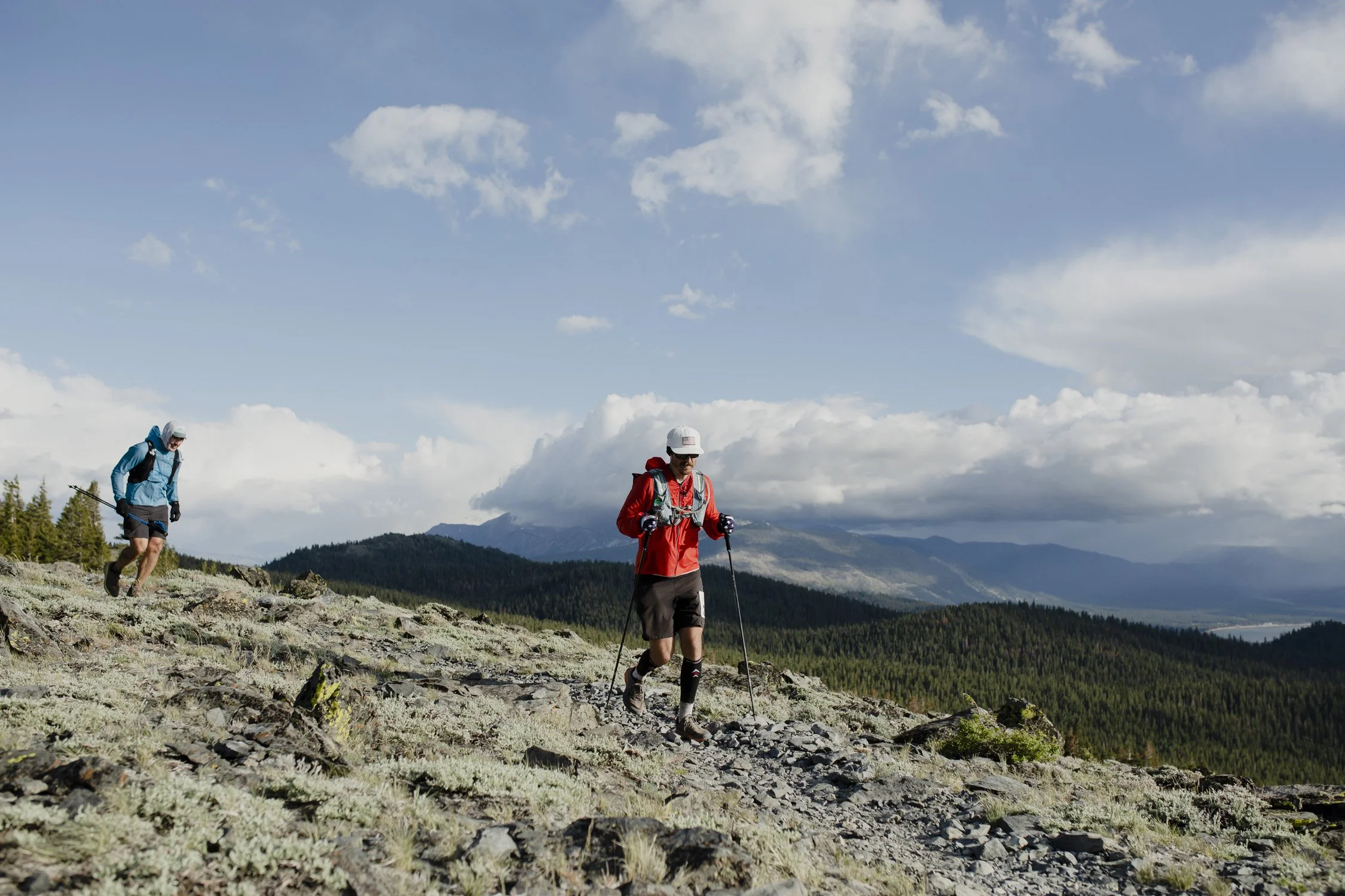 Two hikers trekking on a rocky mountain trail with a forest and mountains in the background, under a partly cloudy sky.
