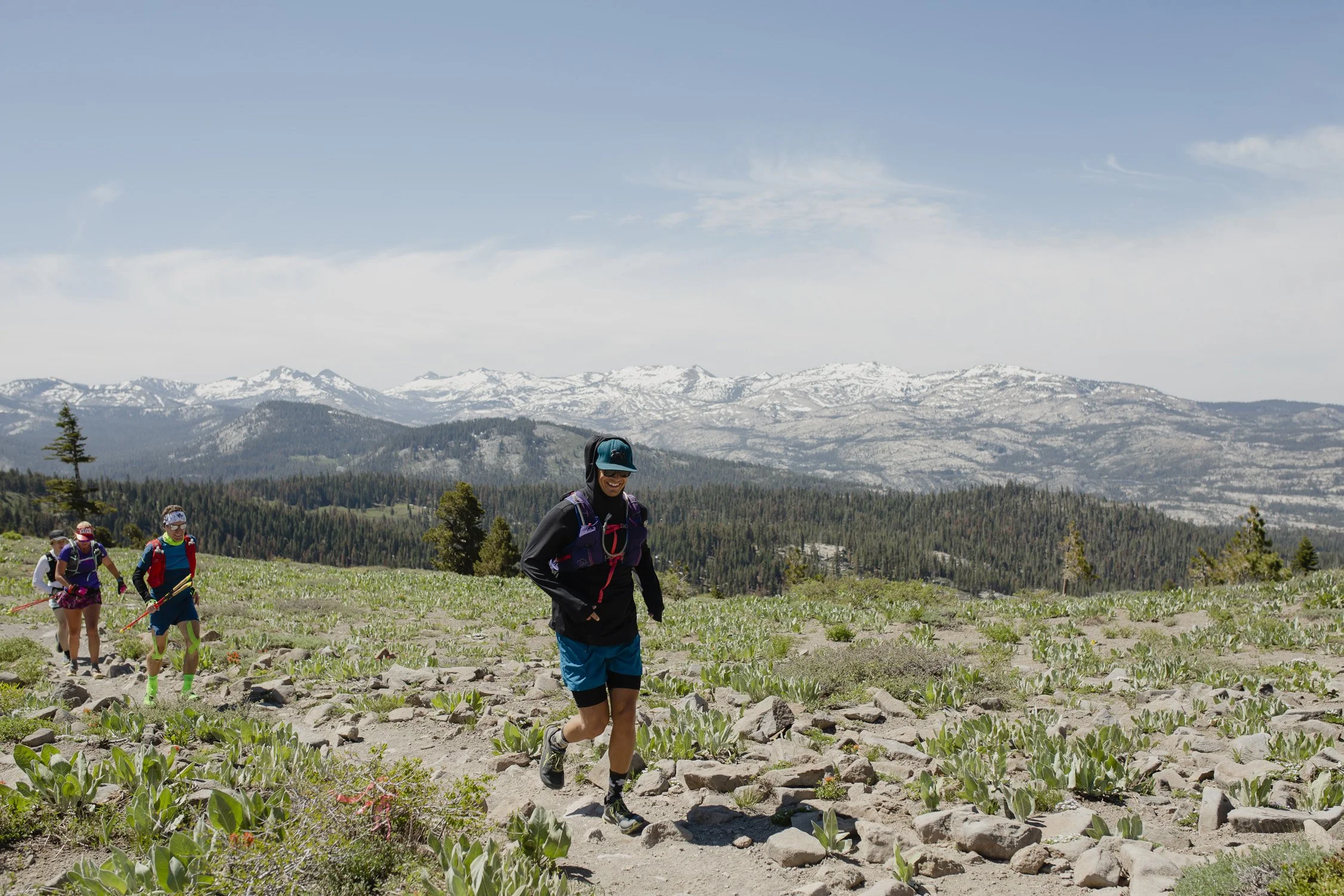 Four trail runners, wearing athletic gear and backpacks, running on a rocky mountain trail with snow-capped mountains in the background.