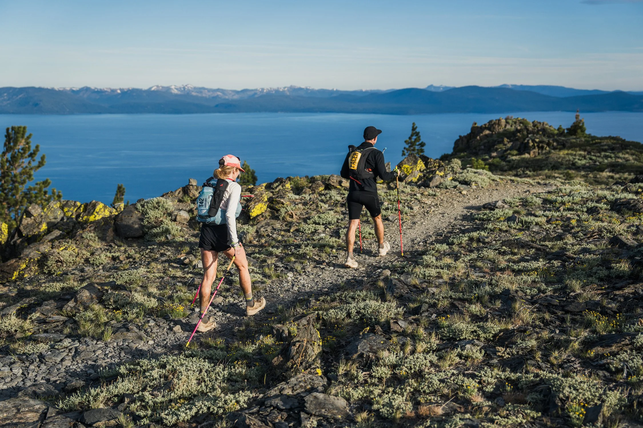 Two hikers, one woman and one man, trekking on a rocky trail with a large lake and mountains in the background.
