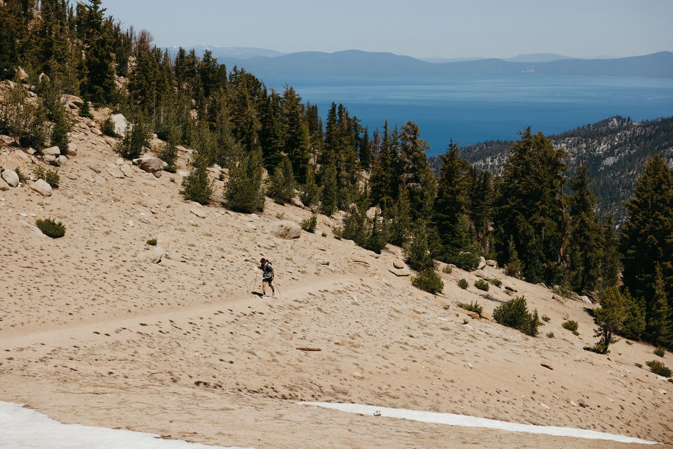 Hiker ascending a mountain trail with trees and snow patches, overlooking a lake and distant mountains.