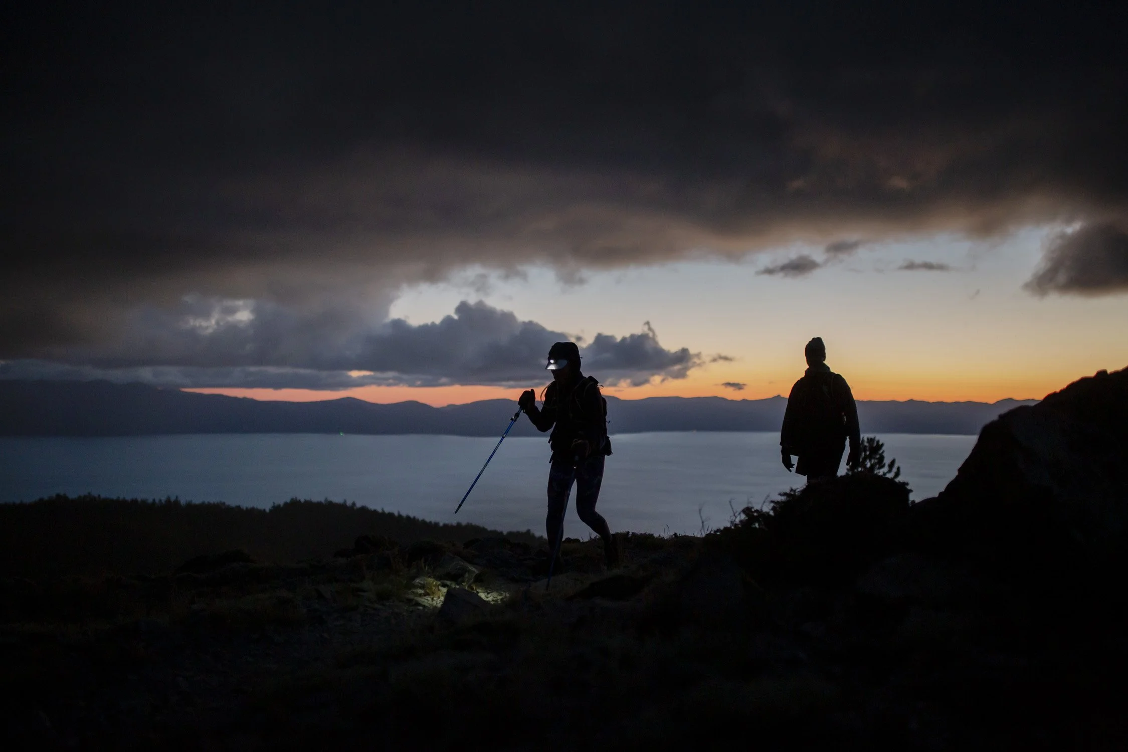 Two hikers silhouetted against a sunset sky, with dark clouds overhead, walking on a rocky trail near a body of water.