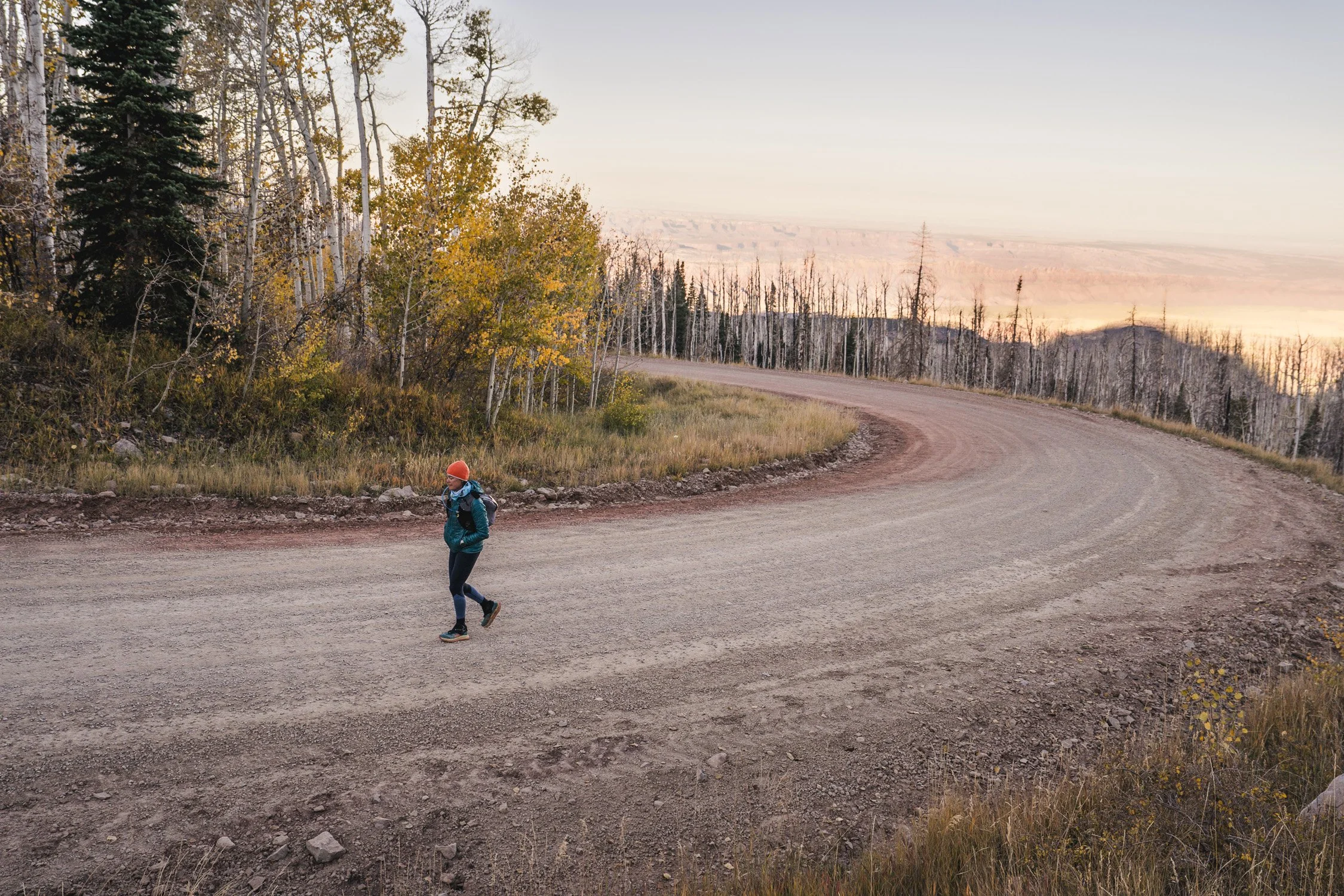 A person hiking on a curved dirt road in a forested area during sunset, with autumn trees and mountains in the background.