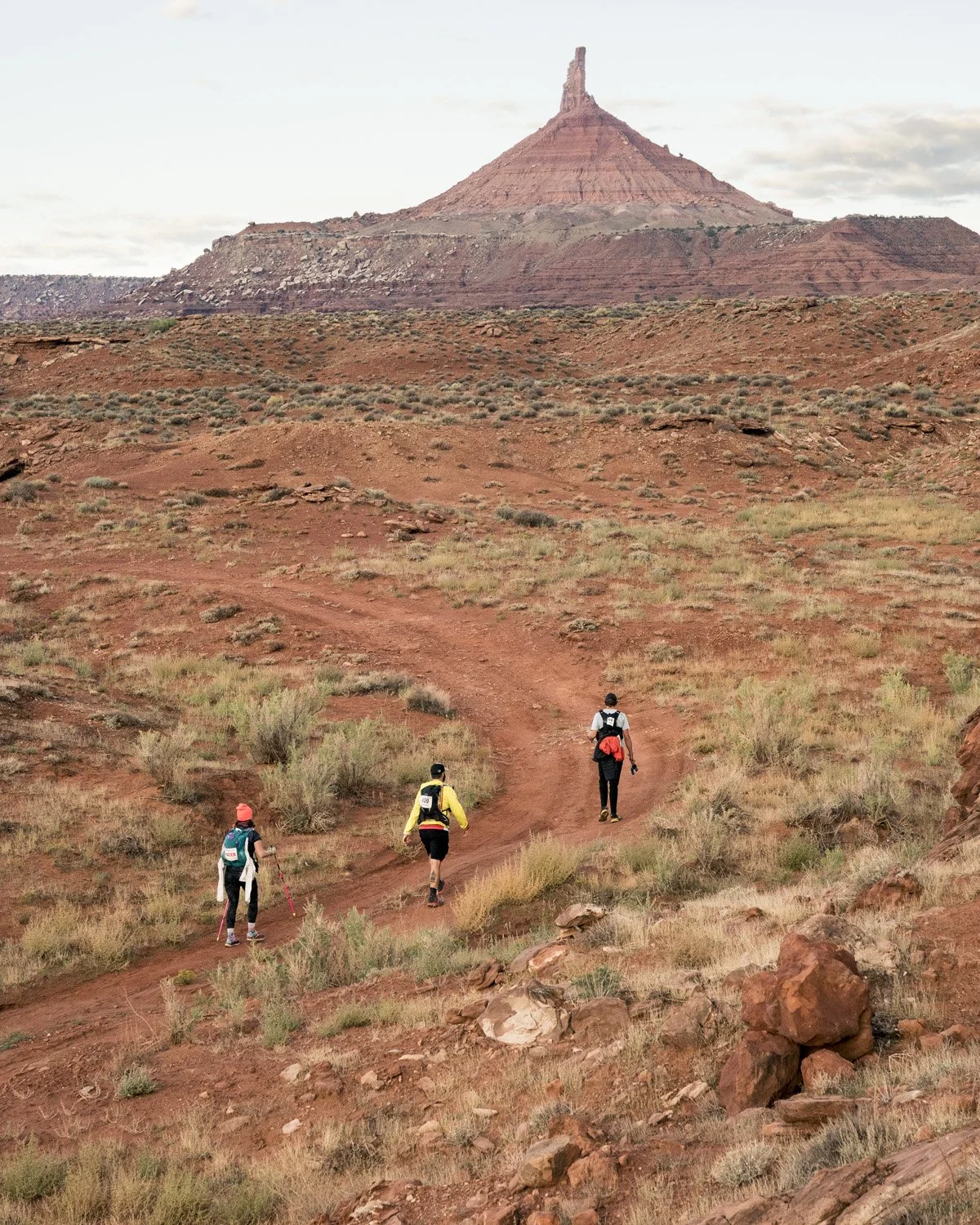 Three hikers walking on a dirt trail through a desert landscape with rocky terrain and sparse vegetation, with a large mesa or butte in the background under a cloudy sky.