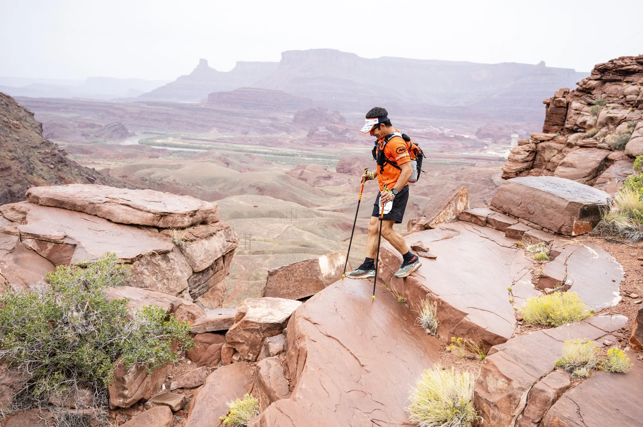 A man hiking on red rocky terrain in a desert landscape, holding hiking poles, wearing an orange shirt, black shorts, and a backpack.