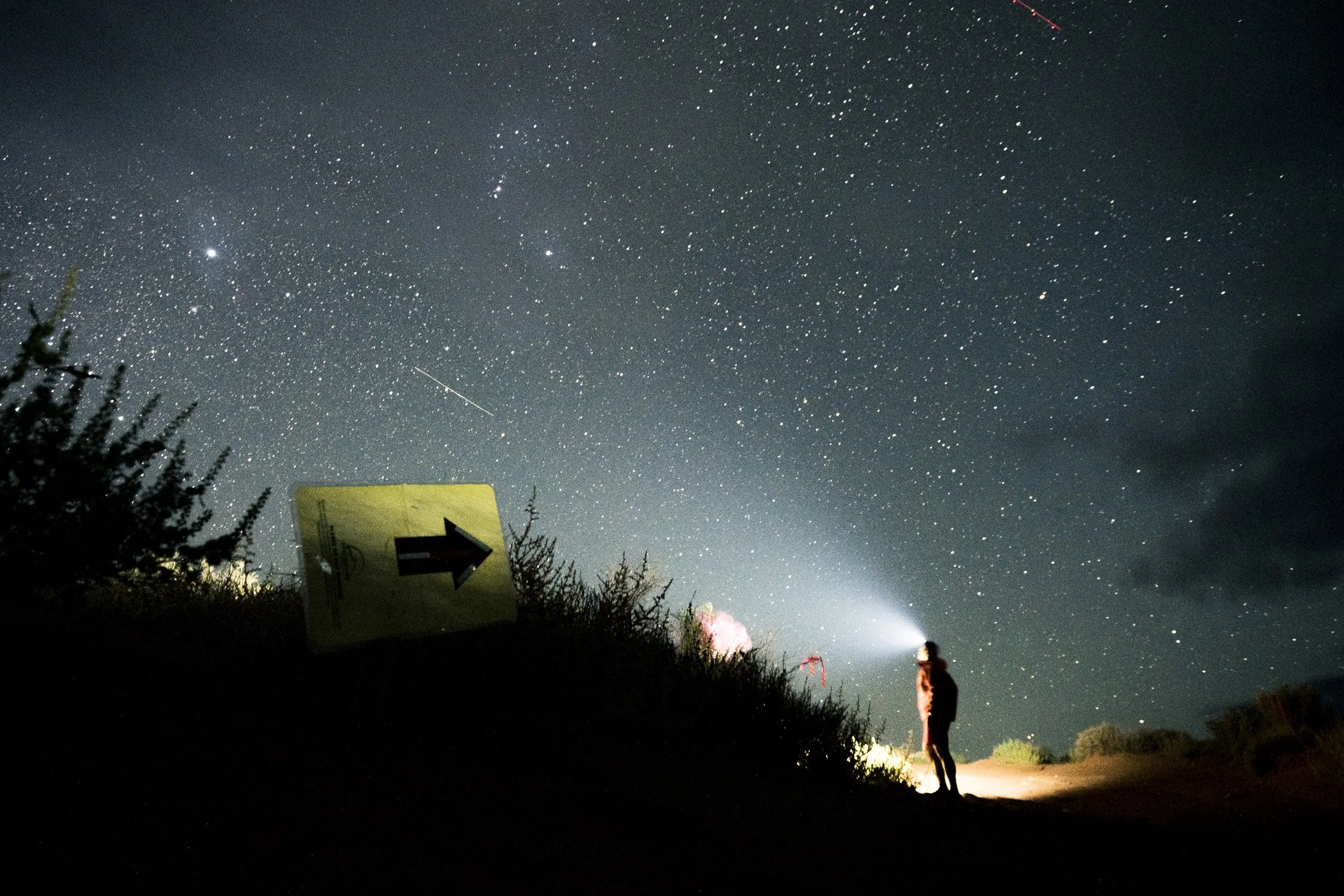 Person standing on a dirt path at night with a headlamp, looking up at a star-filled sky and shooting stars, with a yellow sign pointing right.