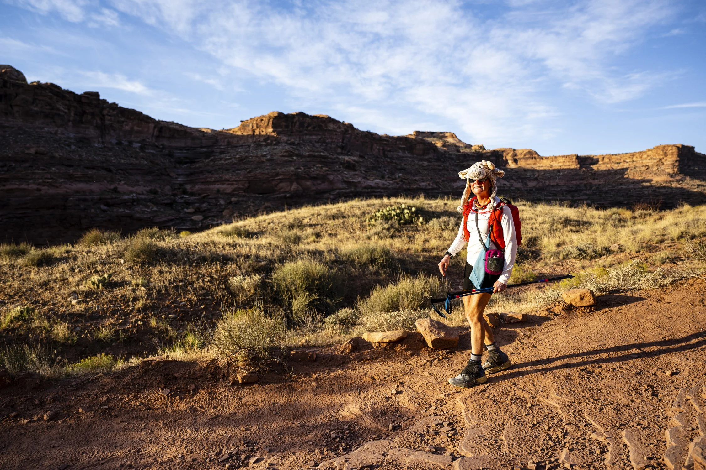A woman hiking on a dirt trail in a desert landscape with rocky cliffs in the background, wearing a hat with a plush animal, sunglasses, a red backpack, and holding a hiking pole.