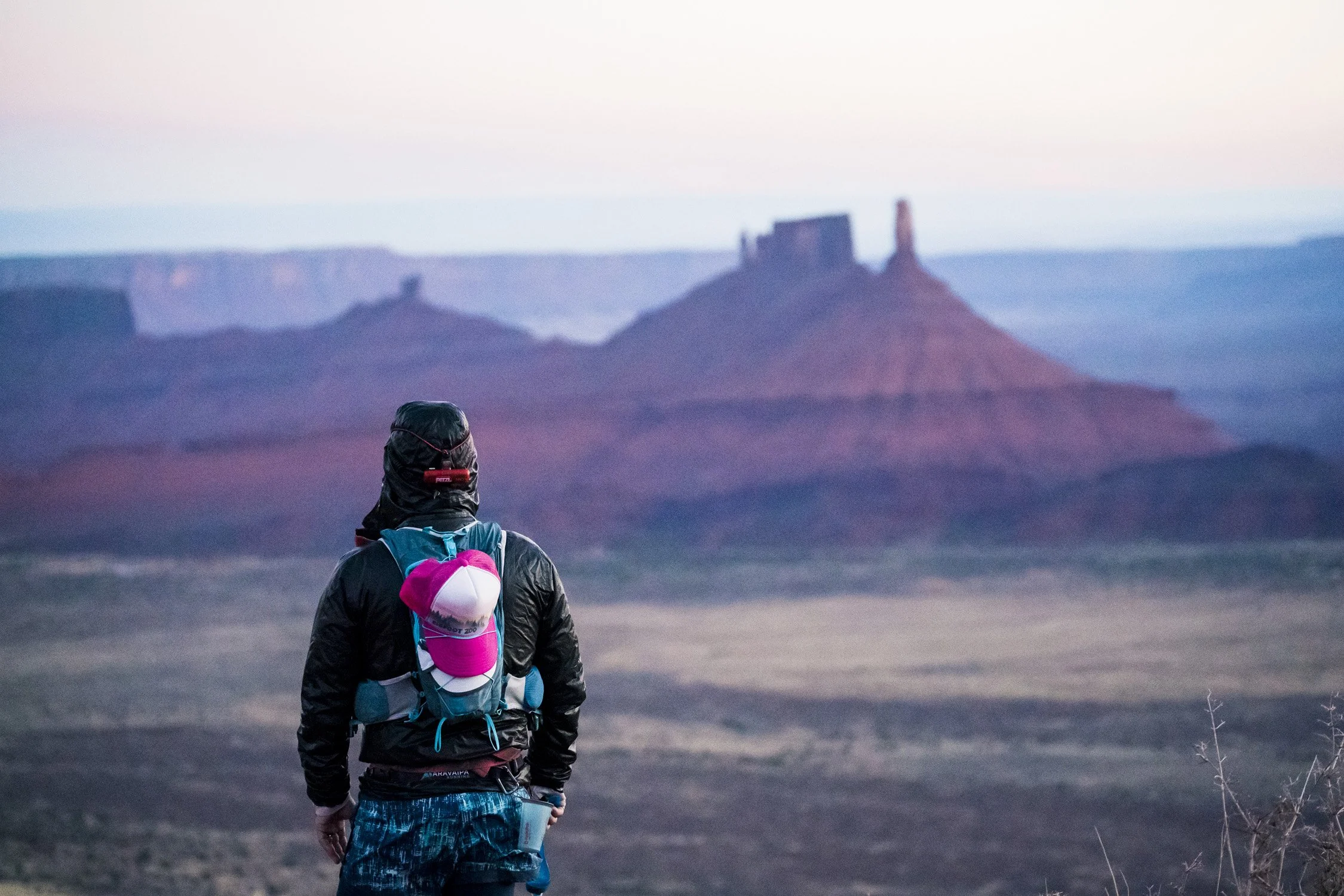 A person wearing a black jacket, blue shorts, and a pink and white cap attached to a backpack walking outdoors in a desert landscape with mesas and buttes in the background during twilight.