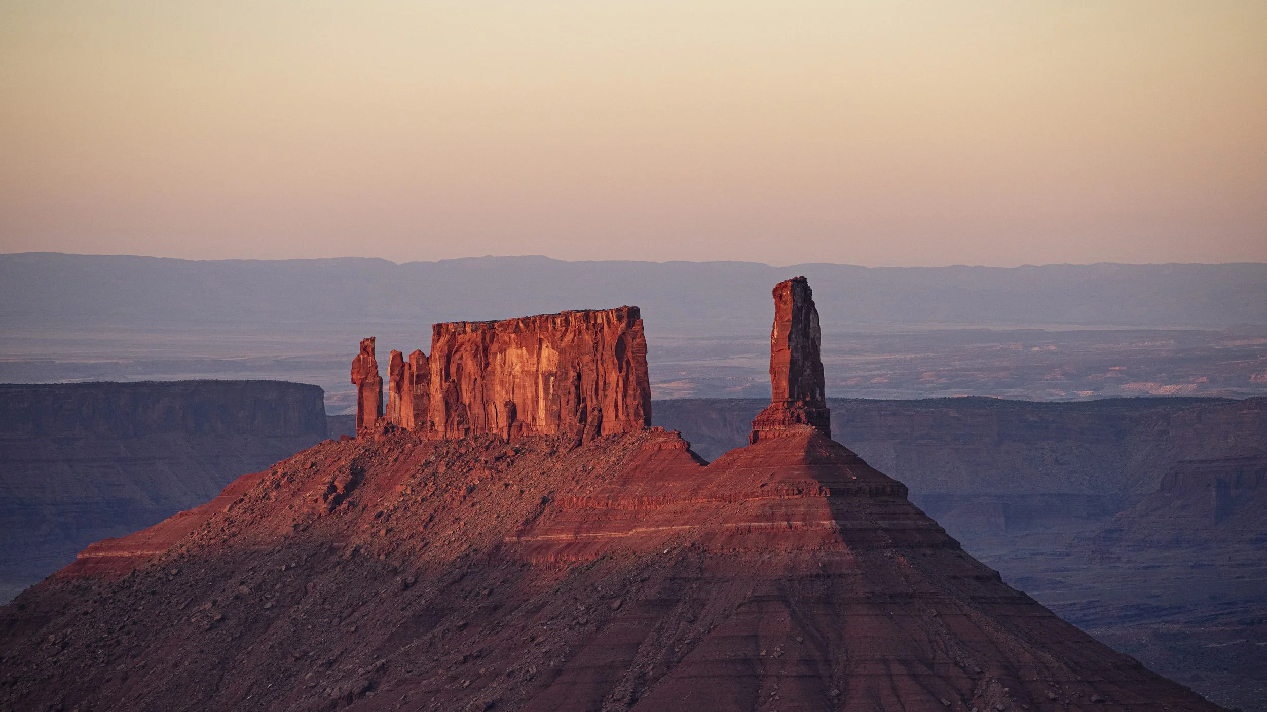 Sunset over Monument Valley with red rock formations, including a flat-topped mesa and towering spires, in a desert landscape.