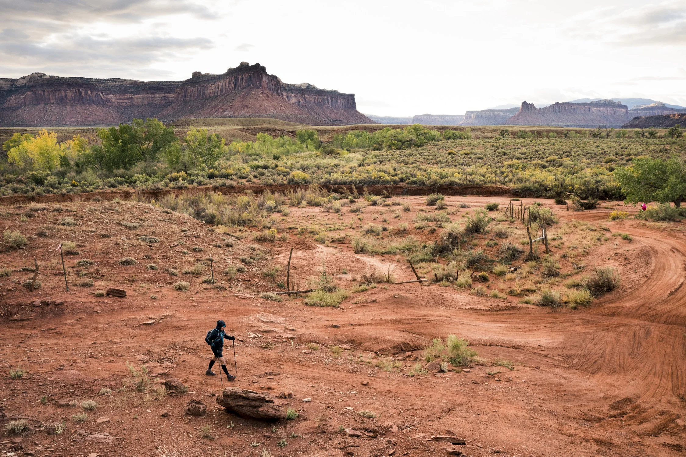 A person hiking with a walking stick on a red dirt trail in a desert landscape, with distant red rock formations and sparse green vegetation under a cloudy sky.