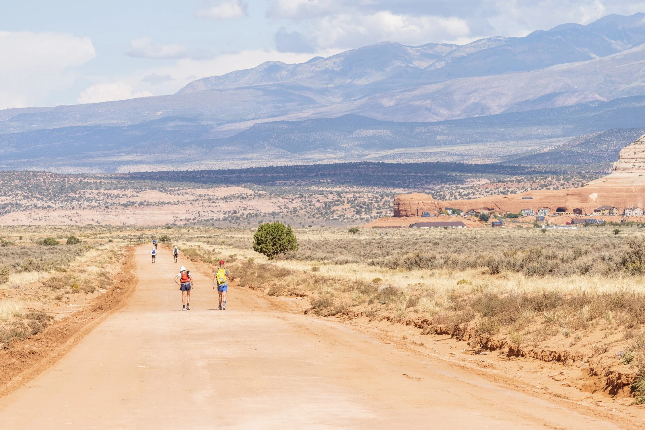 A group of hikers walking along a dirt trail in a desert landscape with mountains in the background and scattered buildings.