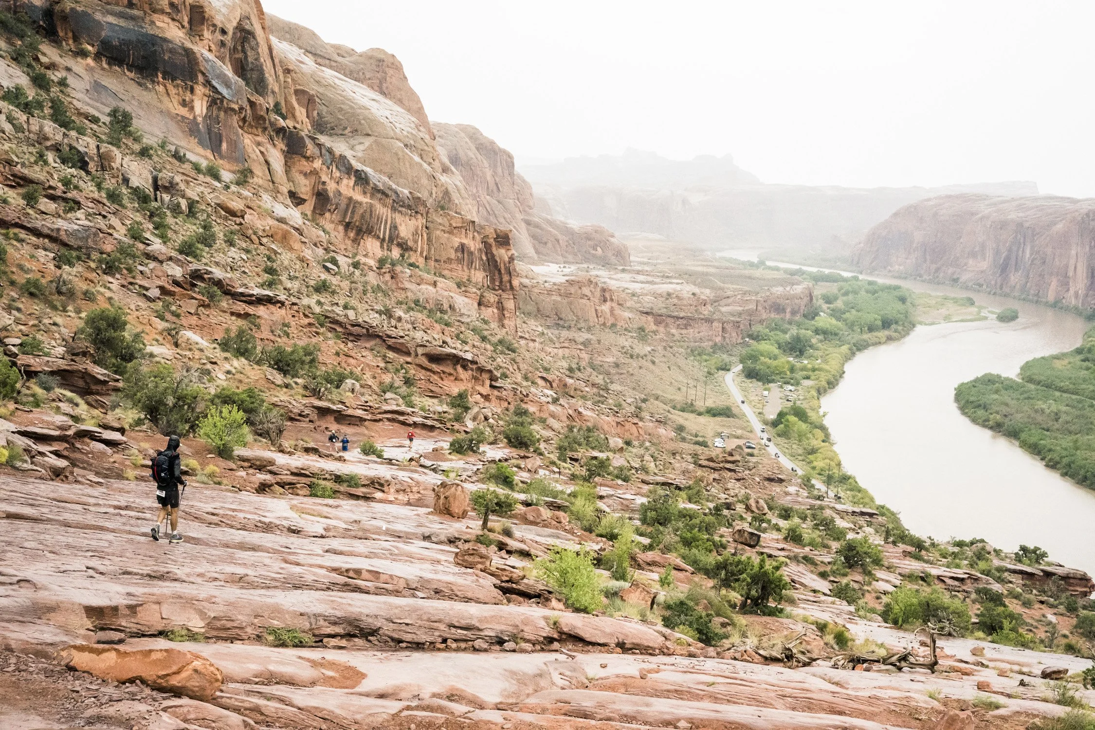 Hiking trail on a red rock slope overlooking a river with lush green trees, cliffs, and a narrow road, under a cloudy sky.