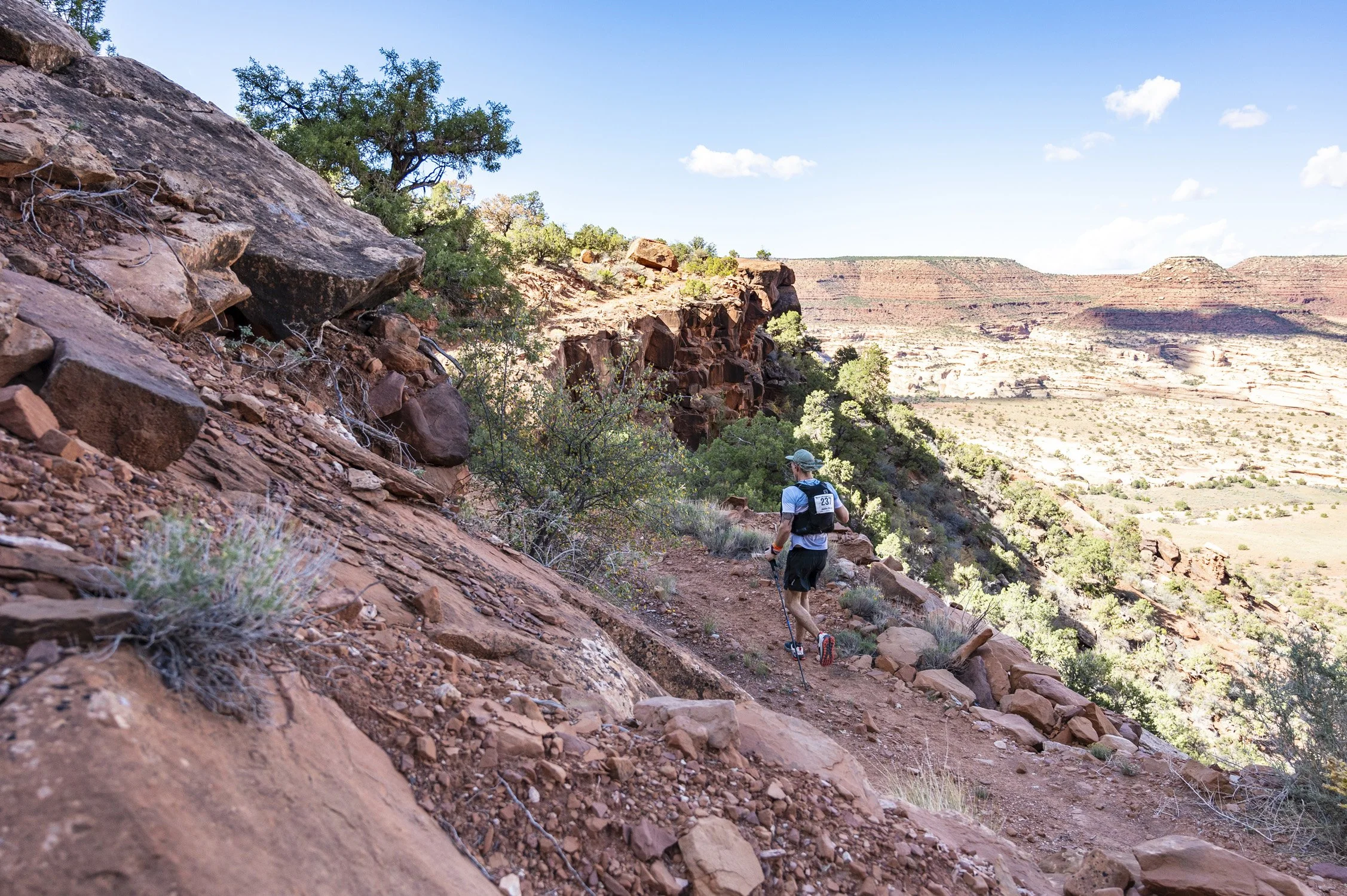 A person trail running on a dirt path through a desert canyon landscape with red rocks, sparse vegetation, and distant mesa formations under a blue sky.