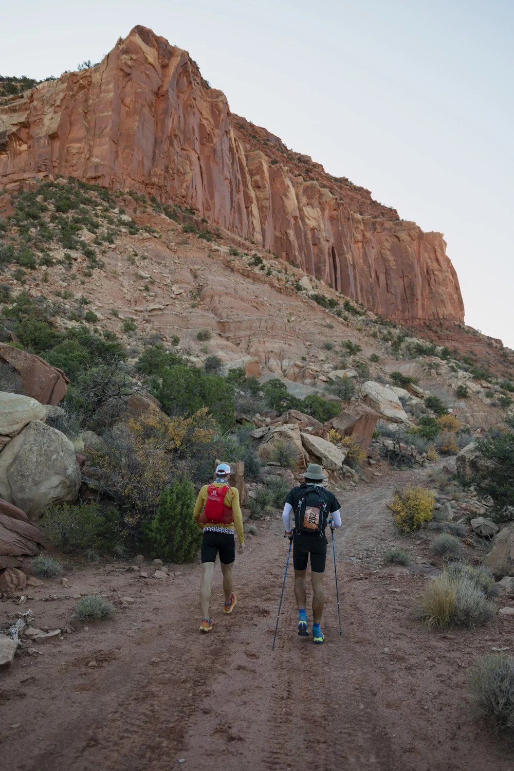 Two hikers walking on a dirt trail in a desert landscape with red rock formations and sparse green and yellow vegetation.