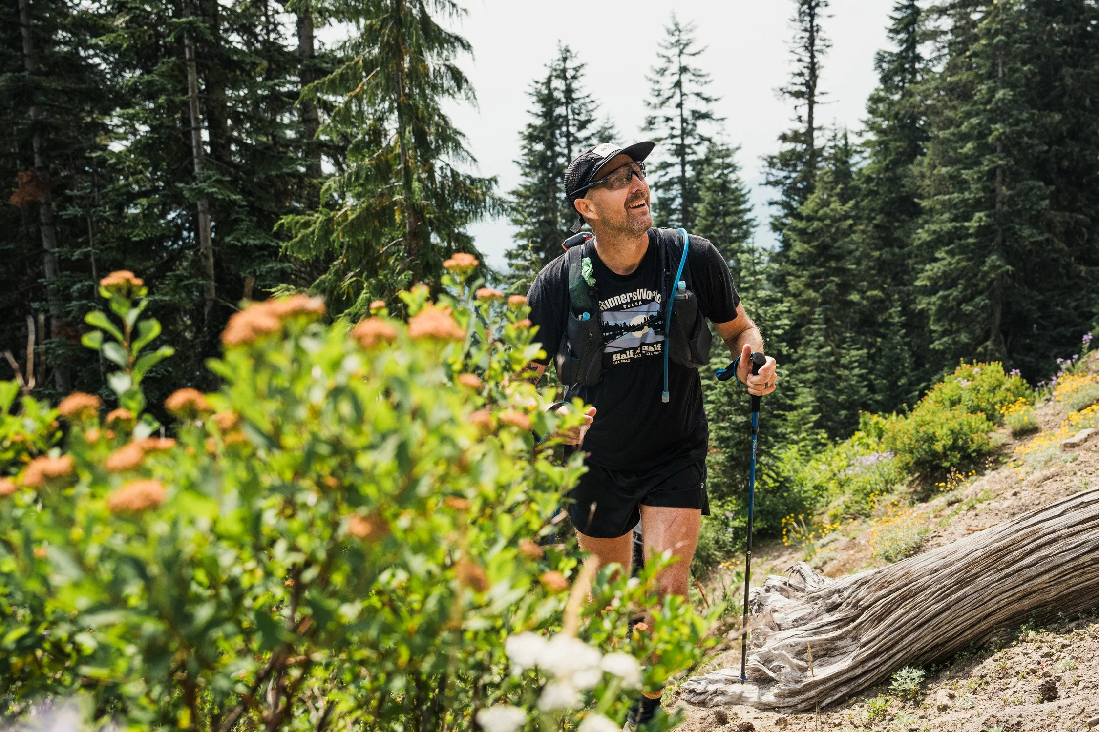 A man trail running on a forest trail surrounded by green trees and bushes, wearing a black t-shirt, shorts, sunglasses, and a cap, using trekking poles.