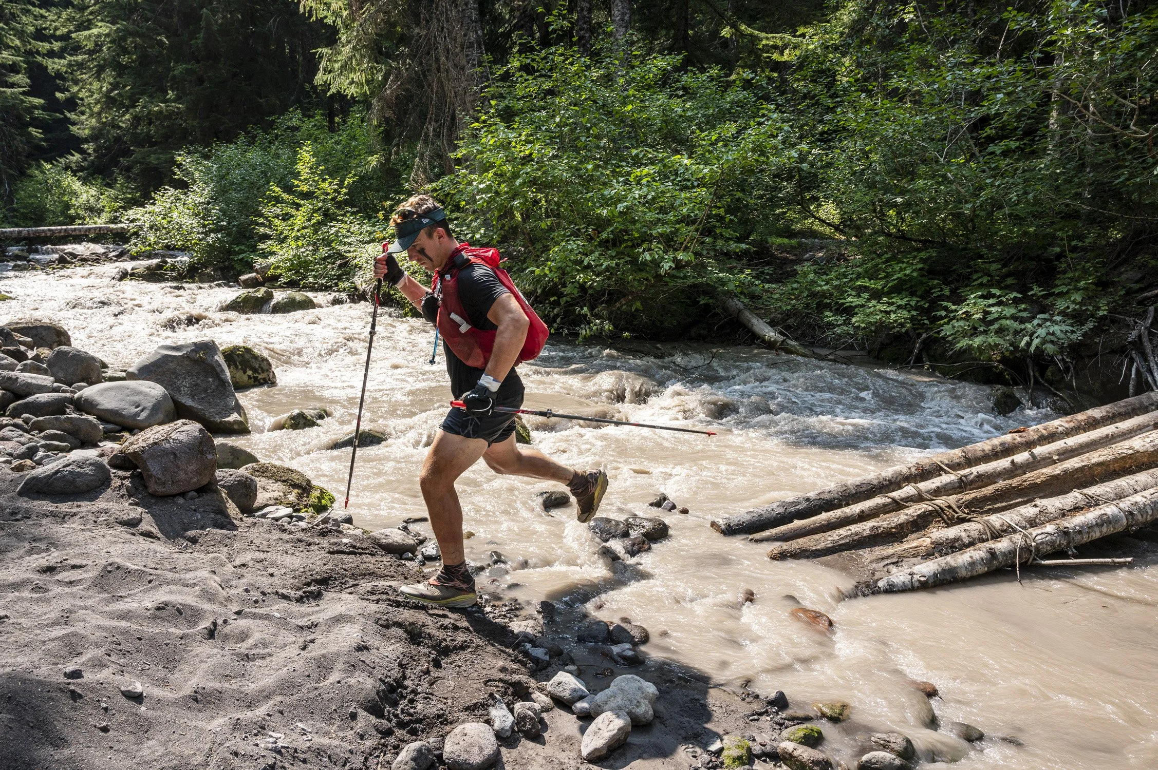 Man wearing black athletic gear and a red backpack crossing a muddy river with rocky banks, surrounded by green trees in a forest