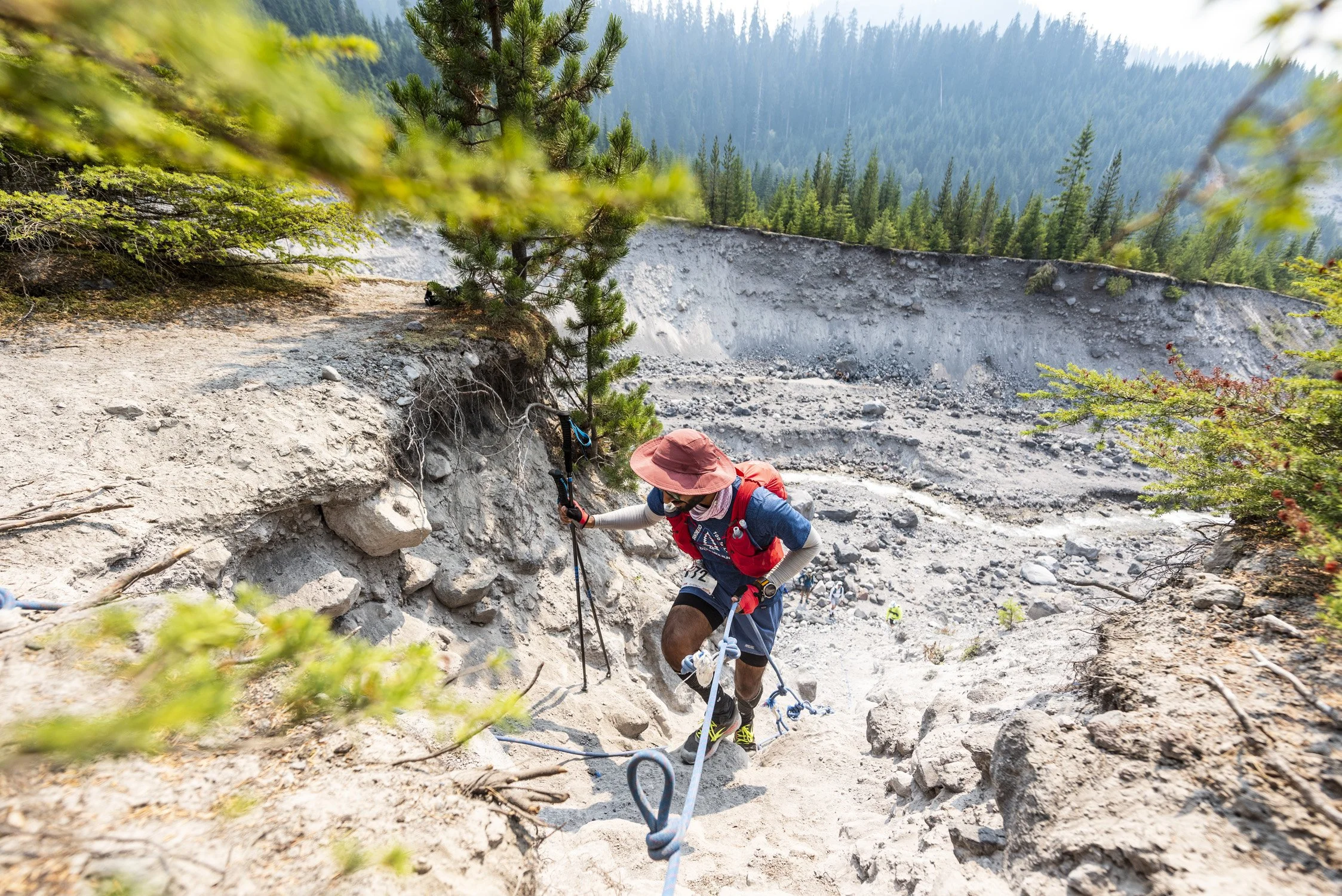 A person wearing a wide-brim hat, sunglasses, and a face mask is climbing up a rocky, steep dirt trail using climbing gear and a rope, with a forested mountain landscape in the background.