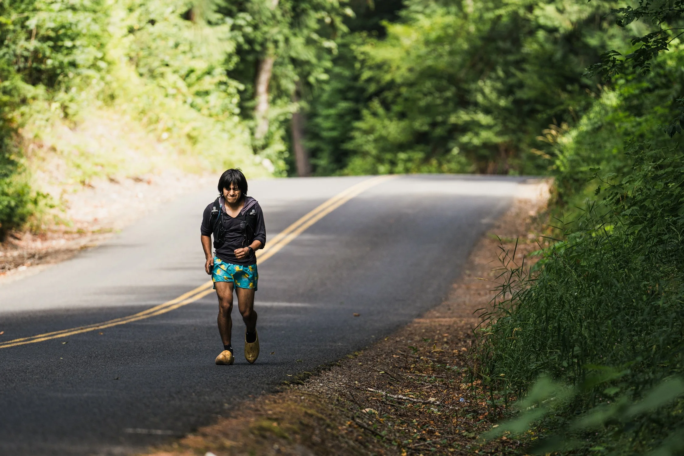 A person running on a curved forest road during daytime, surrounded by green trees.
