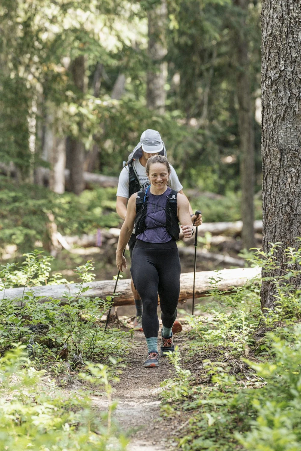 Two women hiking in a forest, using trekking poles and wearing backpacks.