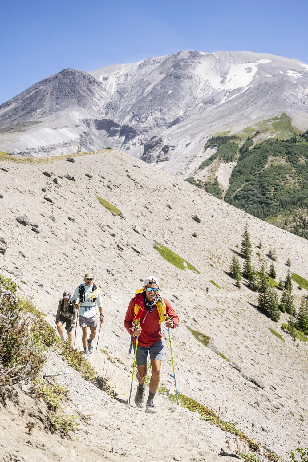 Three hikers trekking up a mountain trail with a snow-capped mountain in the background and greenery along the hillside.