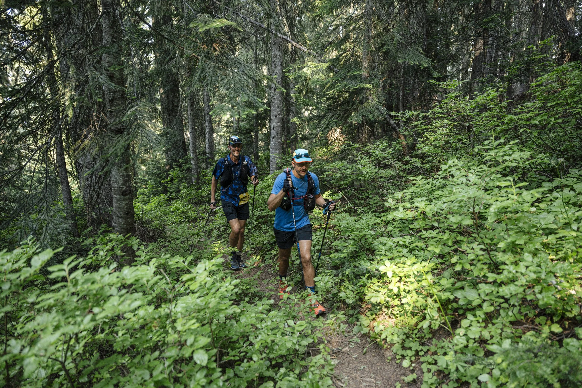 Two men trail running through a lush, green forest with tall trees and dense bushes, wearing athletic clothing, hats, and carrying trekking poles.