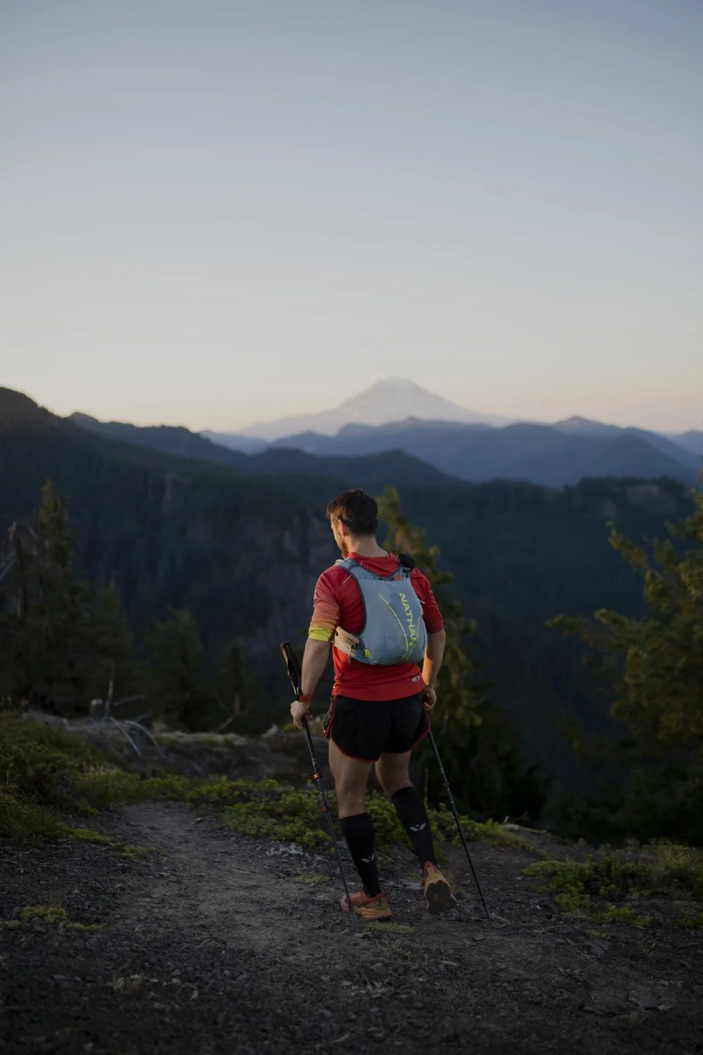 A man hiking on a mountain trail with a backpack and trekking poles, overlooking a forested valley with mountains and a distant snow-capped peak under a clear sky.