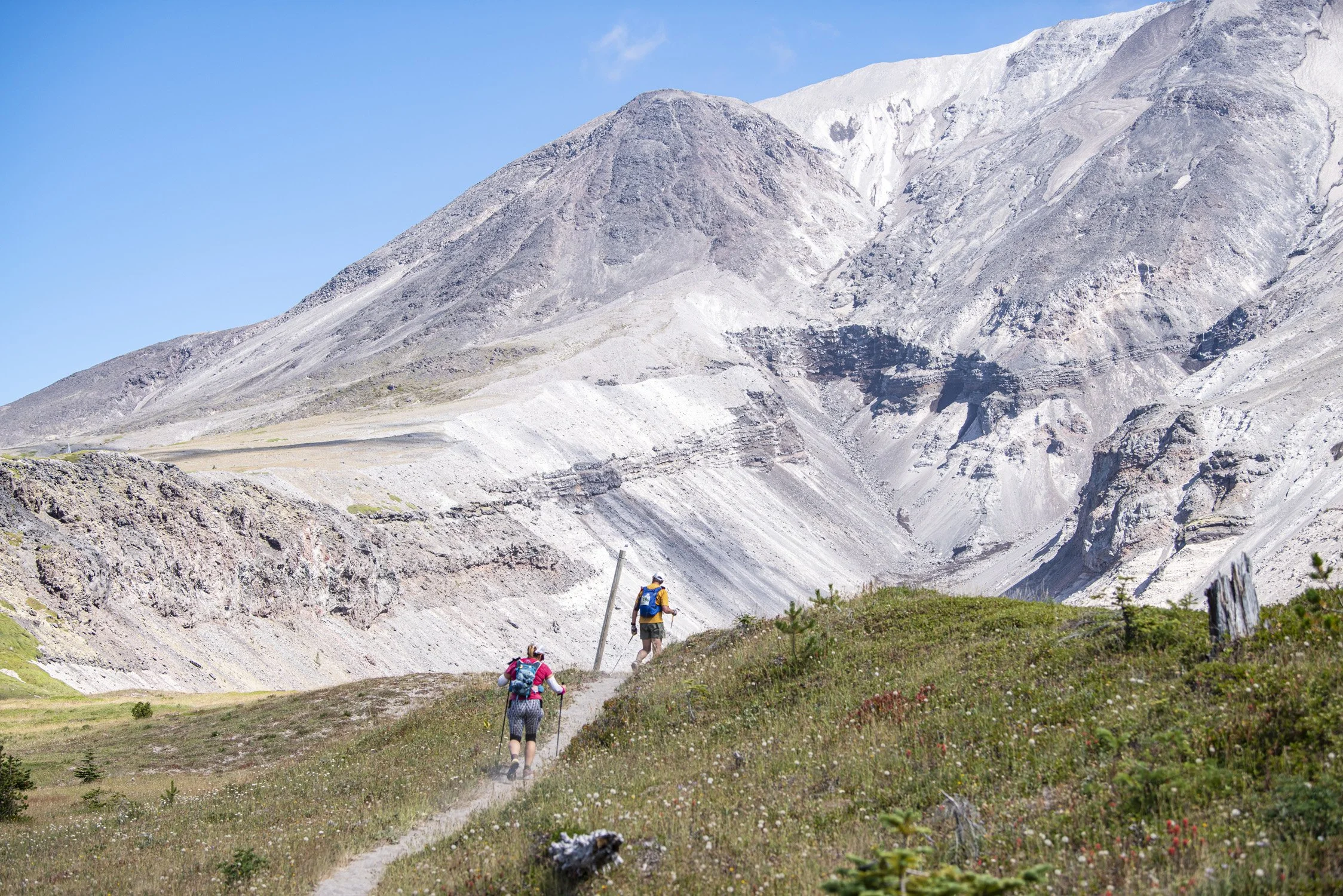 Two hikers walking along a trail in a mountainous landscape with rocky peaks and sparse vegetation.