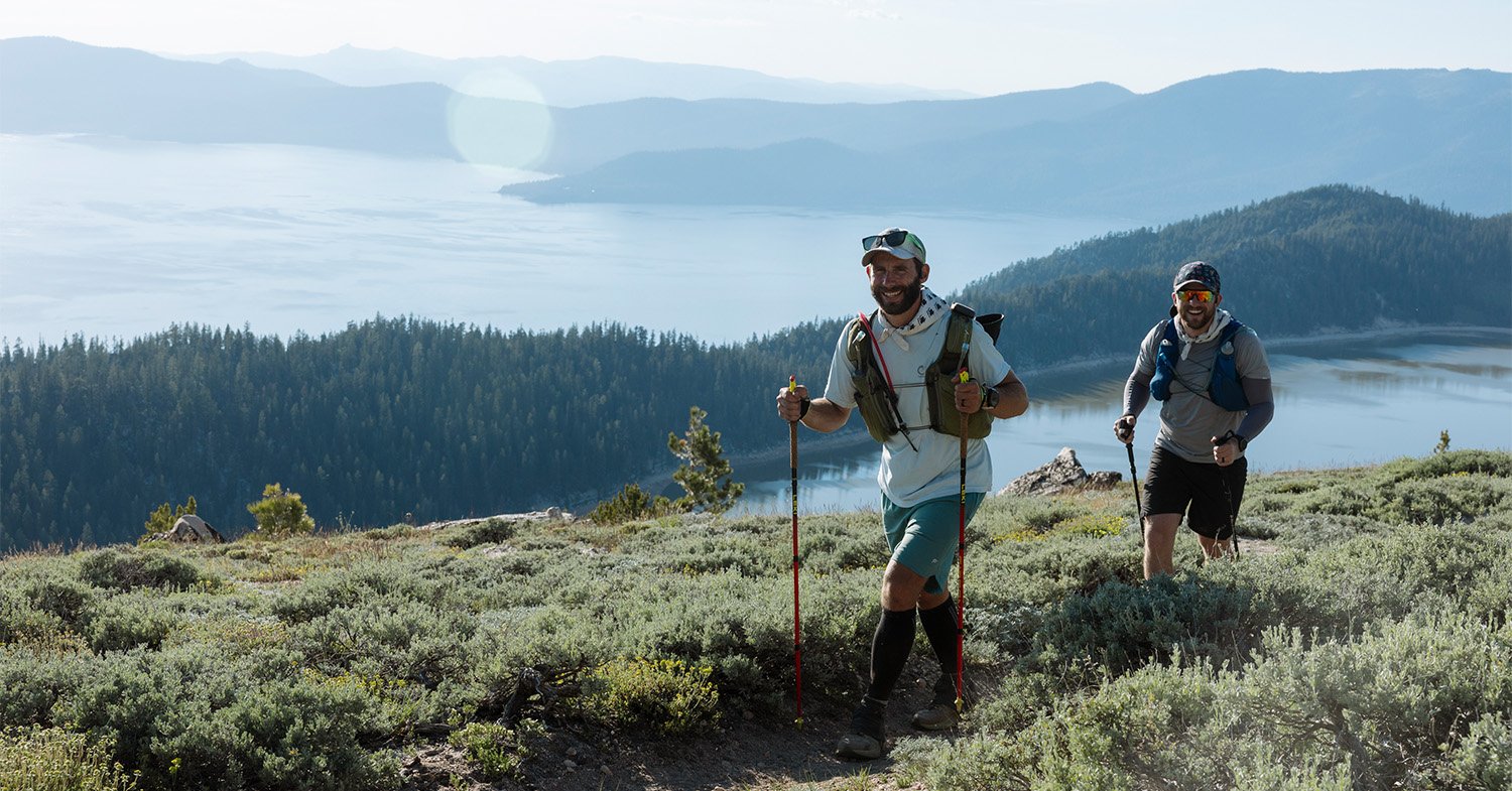 Two runners hike along a trail while smiling with Lake Tahoe in the background
