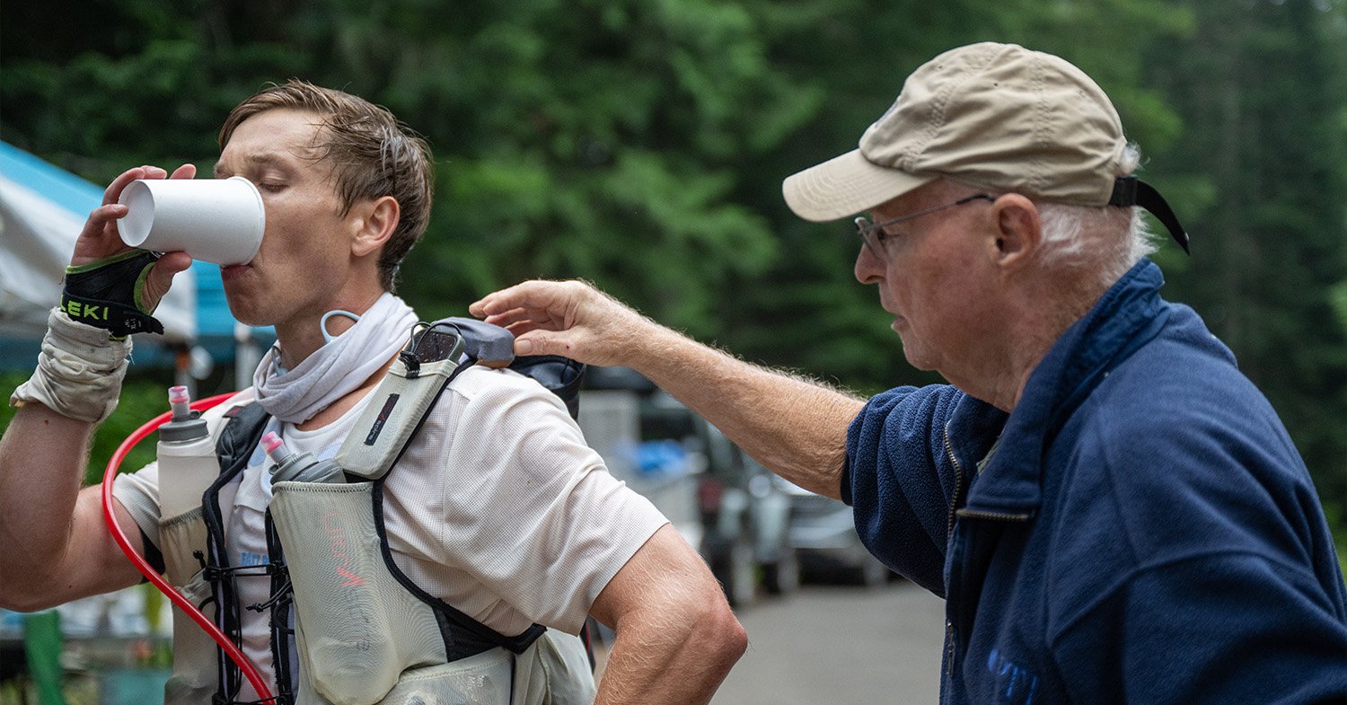 A runner drinks water while a crew member adjusts his running vest