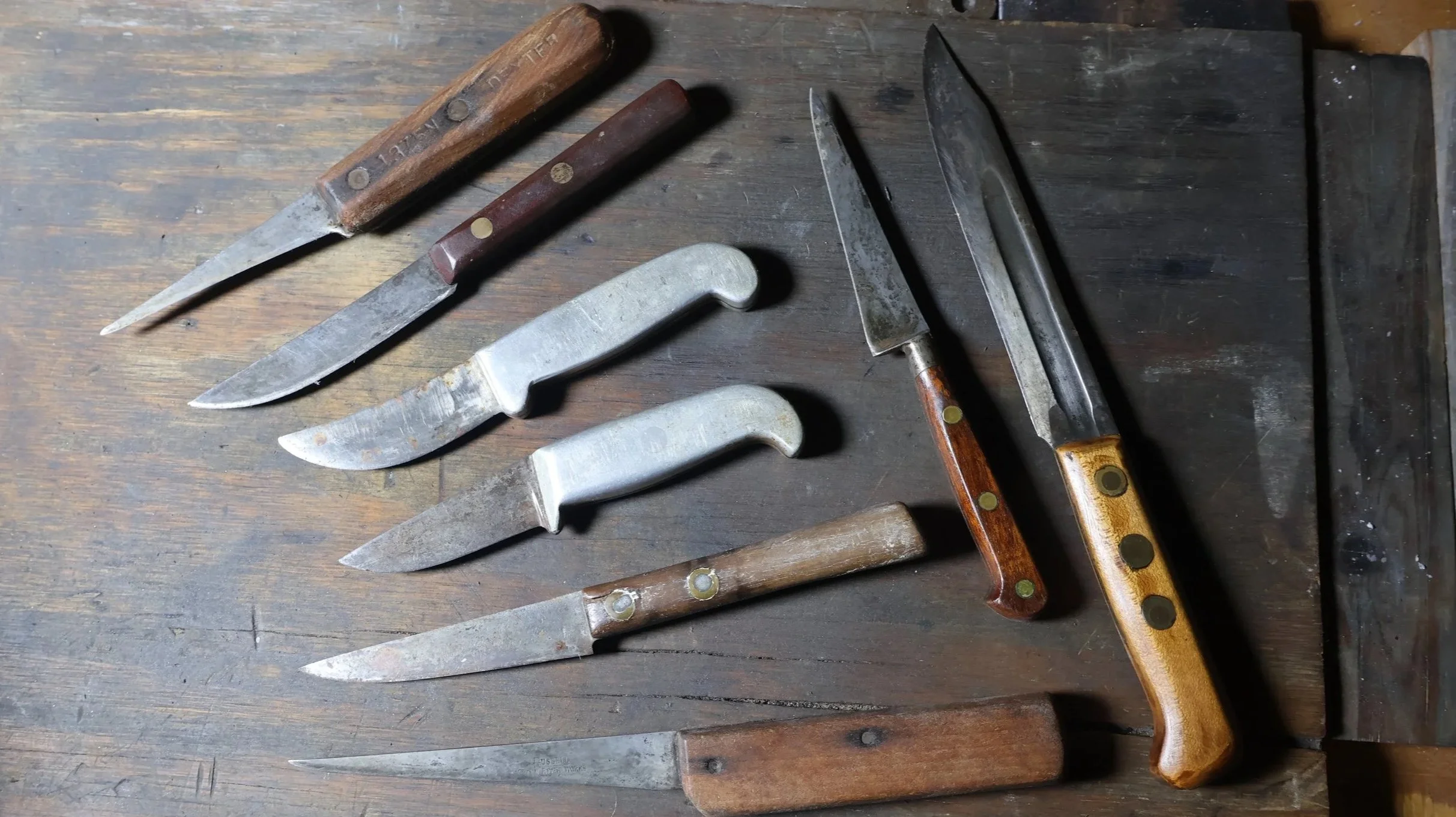 A collection of vintage kitchen knives with wooden, metal, and plastic handles, some with visible rust, lying on a wooden surface next to a sharpening stone. Made by C.K. Chandler.