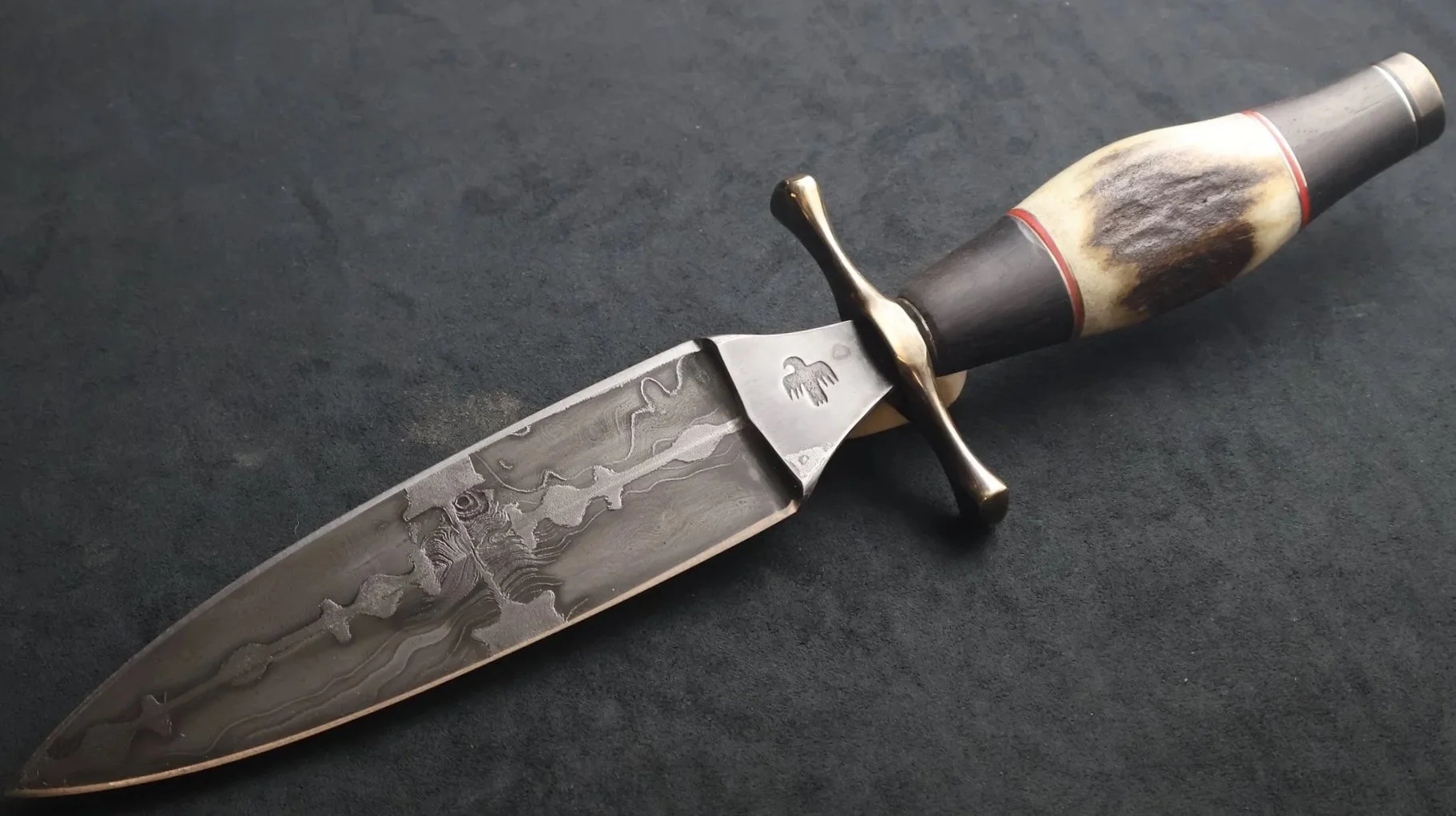 A handcrafted dagger with a Damascus steel blade made from razor blades, an antler handle, and a brass cross-guard, lying on a dark surface. Monty Wheeler - Maker.
