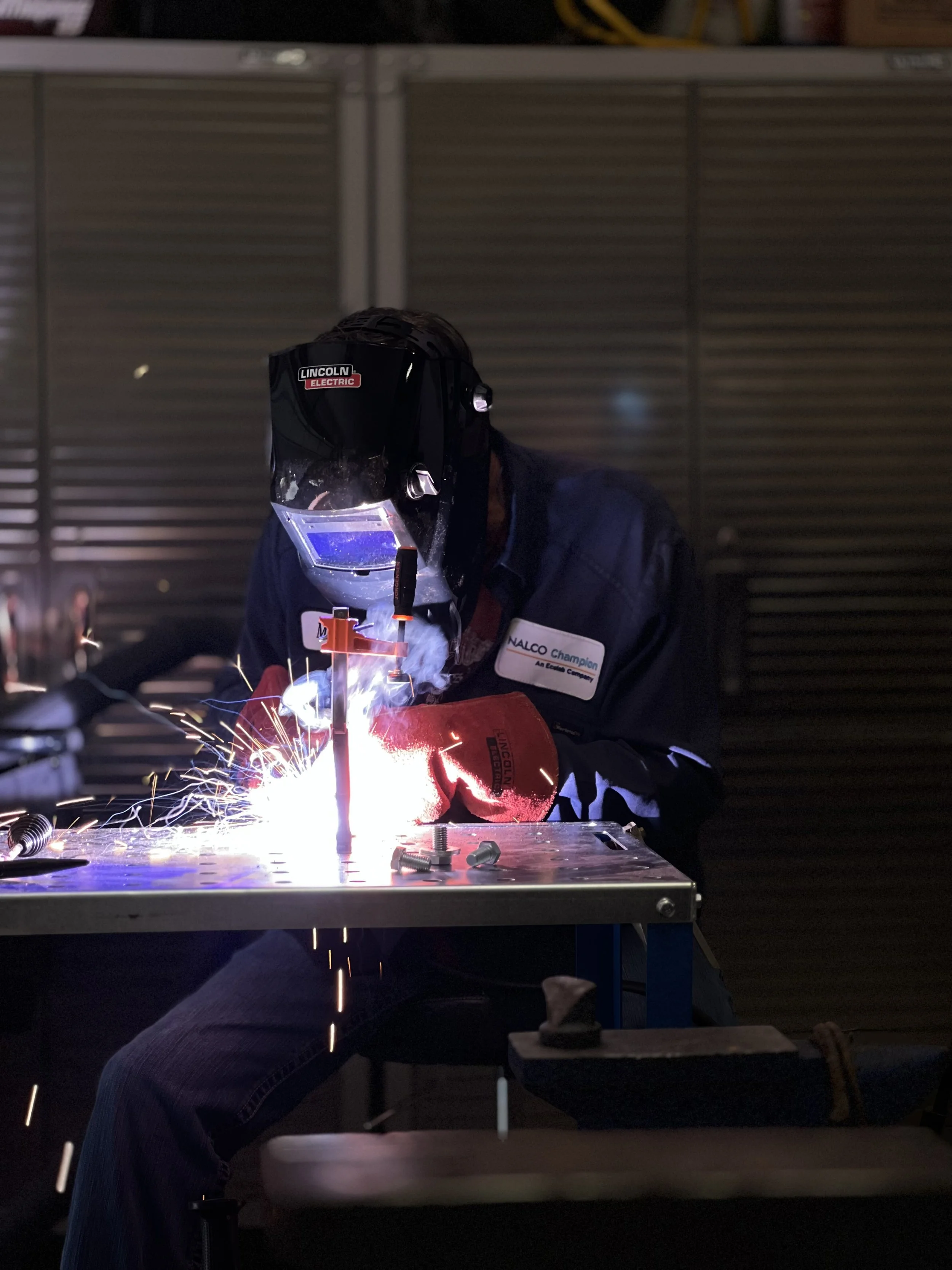 Monty Wheeler welding at a workbench, wearing a protective helmet and gloves, with sparks flying from the welding process.