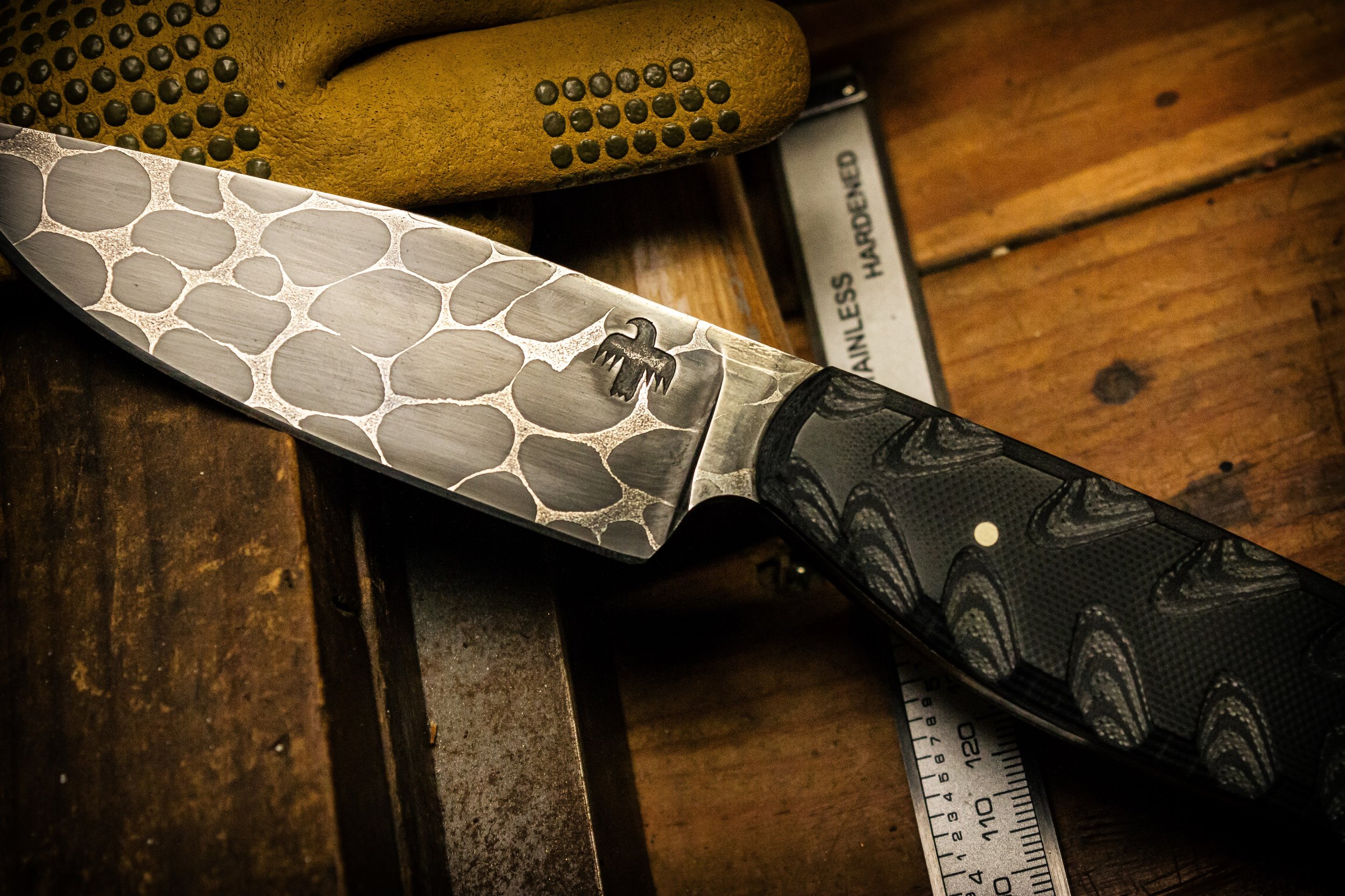 Close-up of a handcrafted knife with a textured black and silver handle and a patterned blade, resting on a wooden workbench. Made by M Wheeler at 66Forge.com.