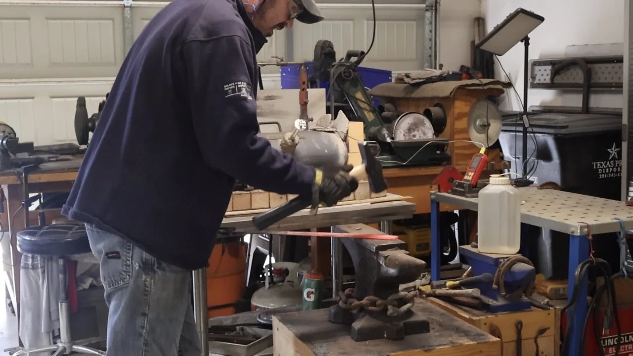 Monty forging in a workshop filled with tools, equipment, and workbenches.