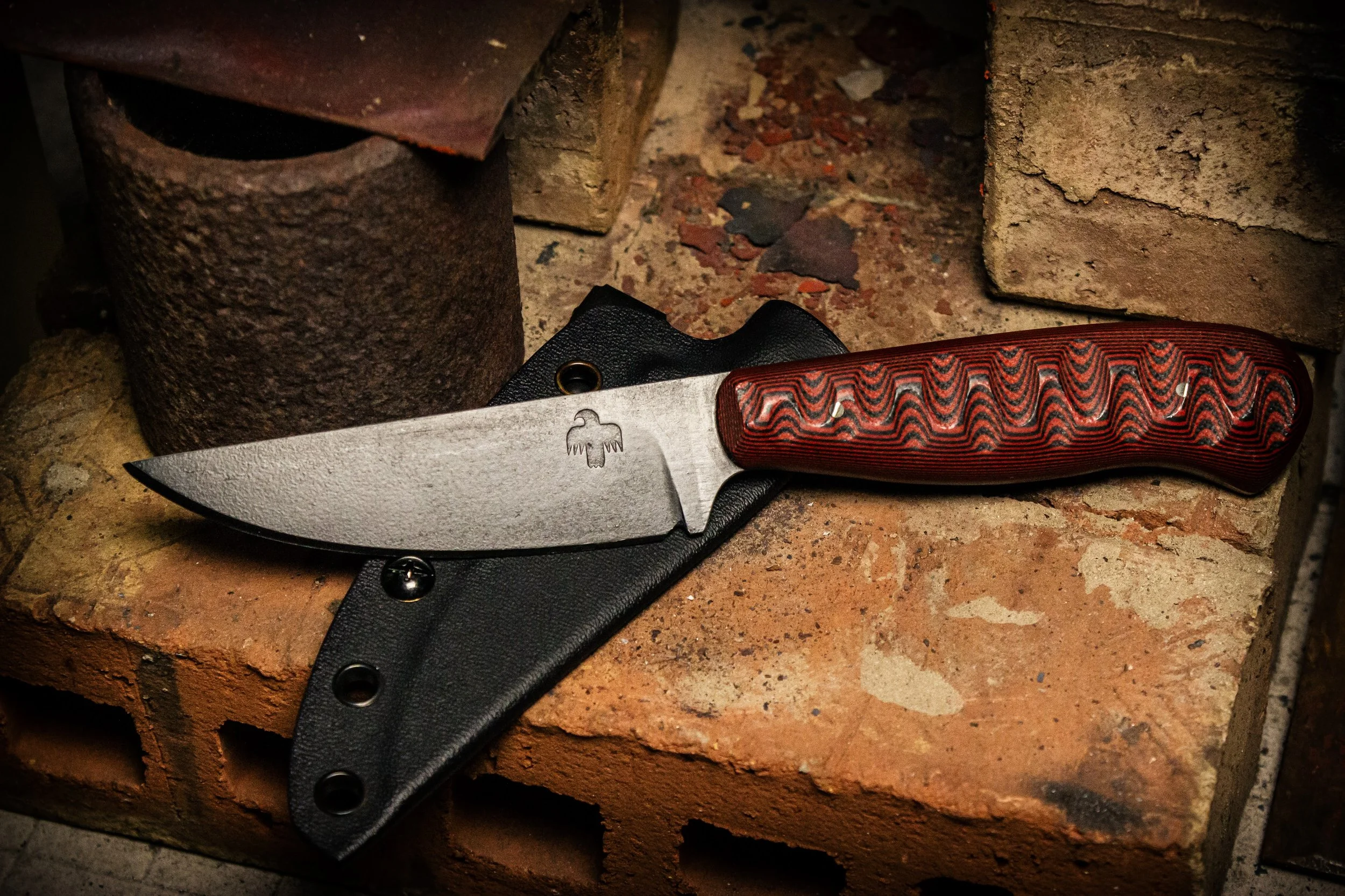 A fixed-blade knife with a textured, red and black handle resting on a black sheath, placed on a brick surface with bricks and rusted metal objects in the background. Made be Monty Wheeler.