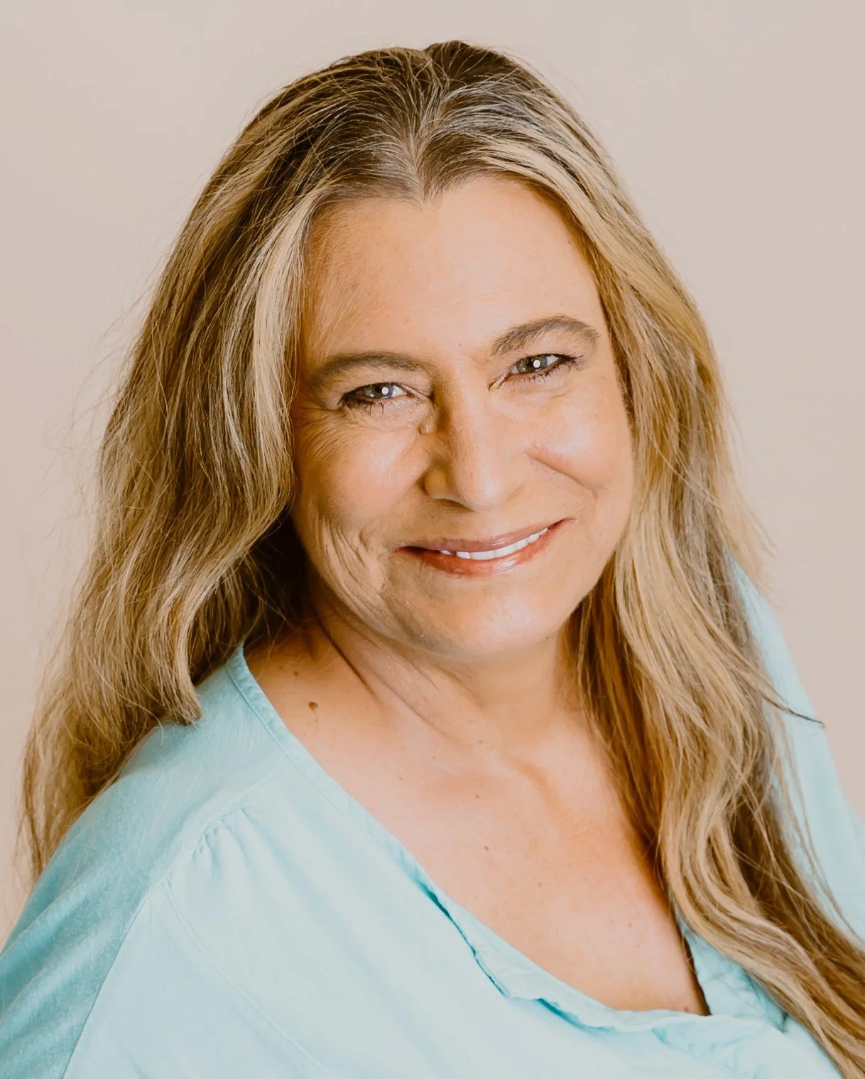 A middle-aged woman with long, wavy blonde hair and blue eyes smiling at the camera, wearing a light blue top against a plain, off-white background.