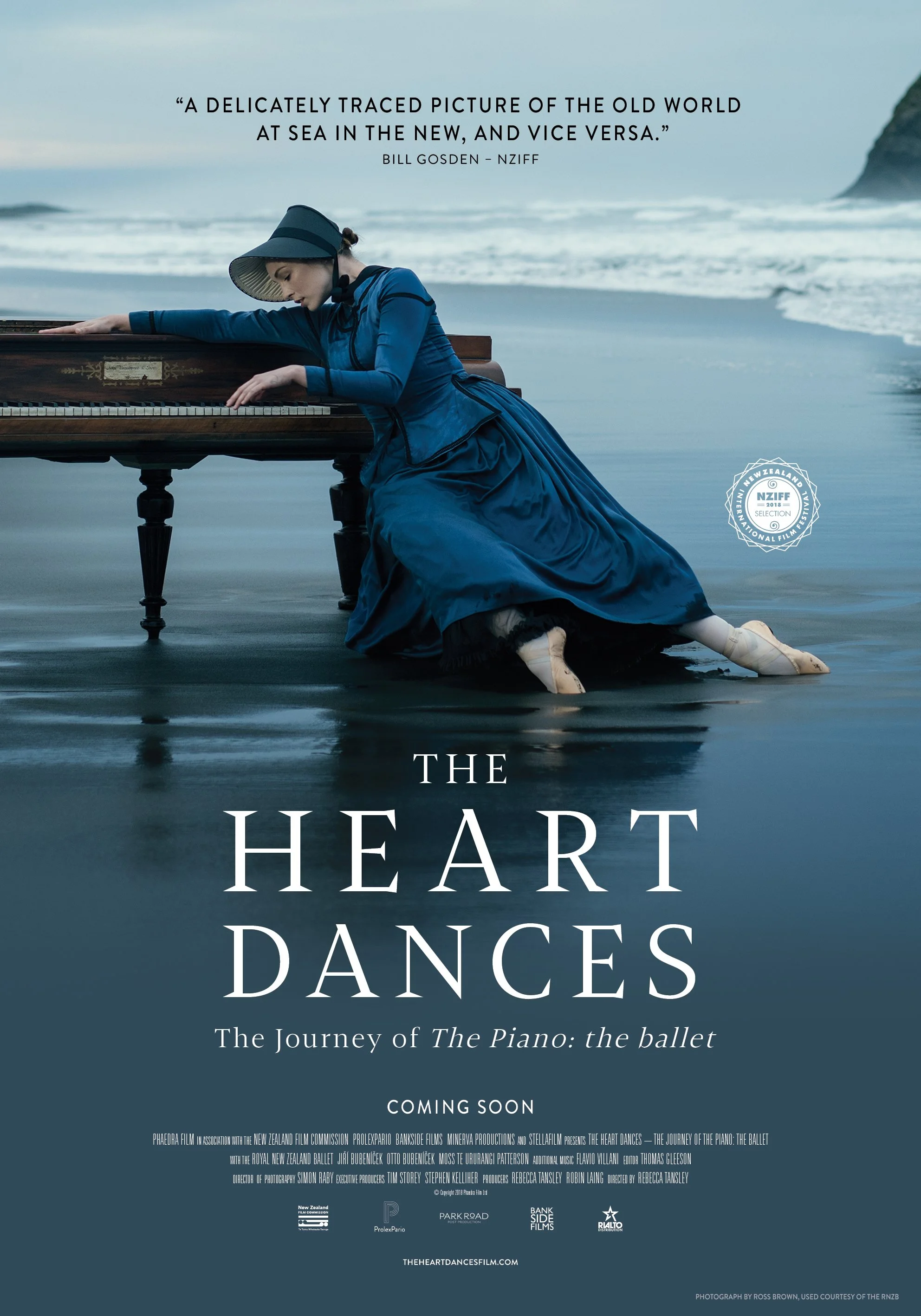 A woman in Victorian-style clothing, dressed in blue, is kneeling on a piano that is partially submerged in water on a beach. She is leaning on the piano with her eyes closed and appears to be resting, with her feet and part of her dress in the water. The background shows the ocean with waves and a rocky coastline.