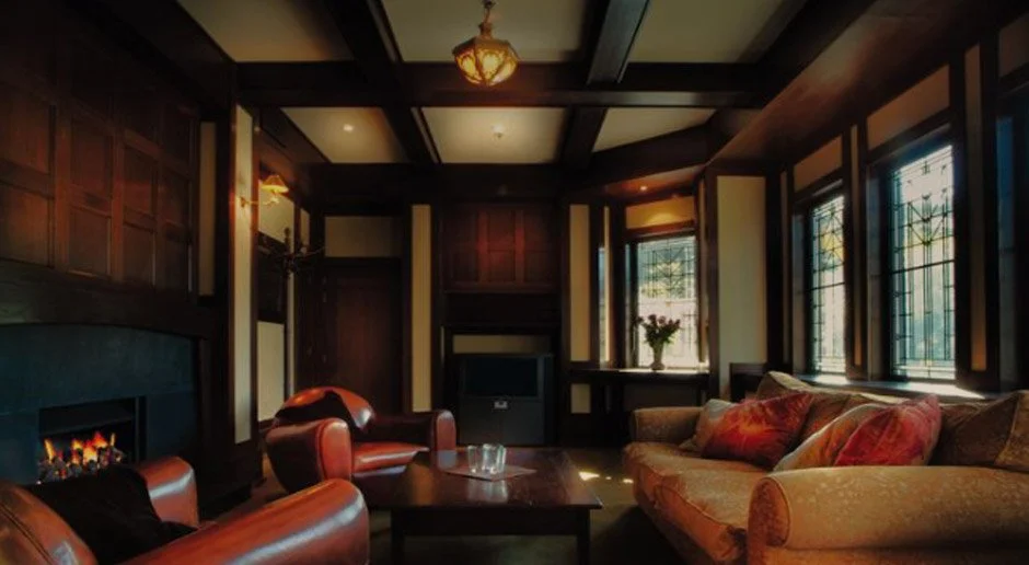 A cozy living room with dark wood paneling, large windows with stained glass, a fireplace, leather armchairs, a coffee table, and a floral arrangement on the windowsill.