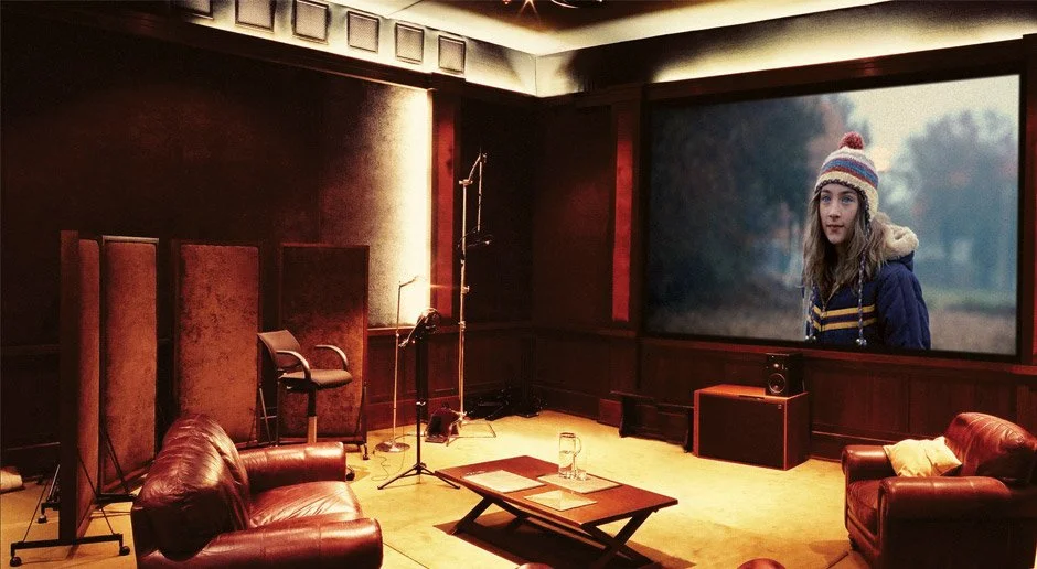 Recording studio with leather sofas, a coffee table, soundproofing panels, a microphone, a music stand, and a large screen showing a girl outdoors wearing a knit cap.