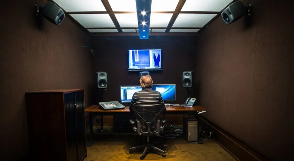 A person working at a computer in a soundproof recording or editing studio, with multiple monitors, speakers, and soundproof walls.