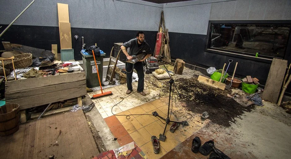 Man using a jackhammer on a floor in a cluttered room with construction tools and materials around.