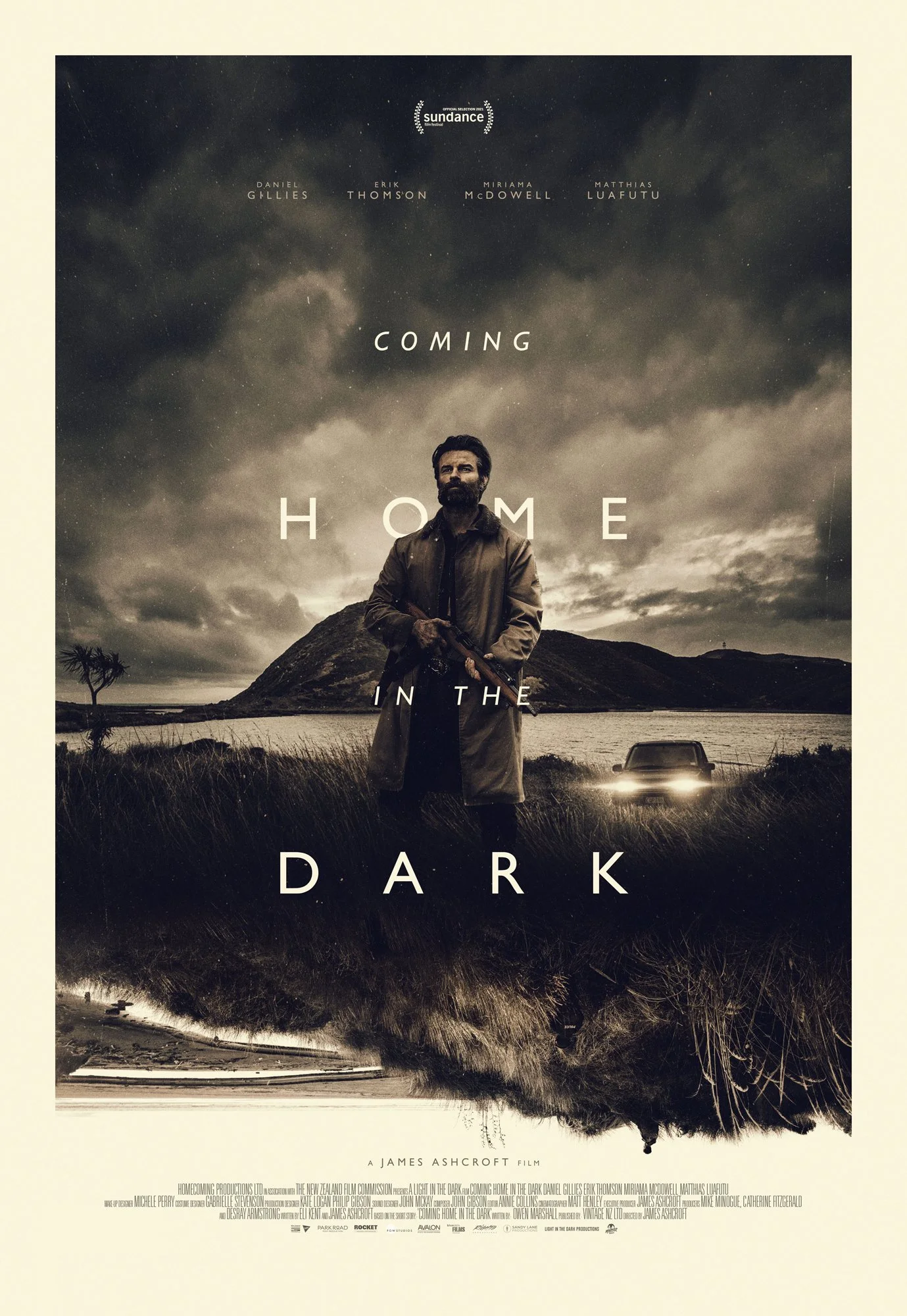 A movie poster for "Coming Home in the Dark" featuring a man with a beard holding a gun, standing near a lake with hills and cloudy skies in the background, and a car with headlights on parked behind him.