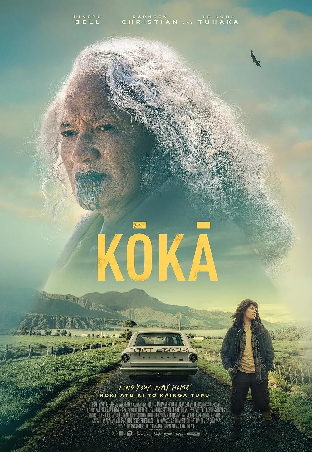 A movie poster for 'Kōkā', featuring an older man with long, curly gray hair and distinctive facial markings, and a young woman standing beside a white car on a rural road with mountains in the background. The sky is partly cloudy with a bird flying.