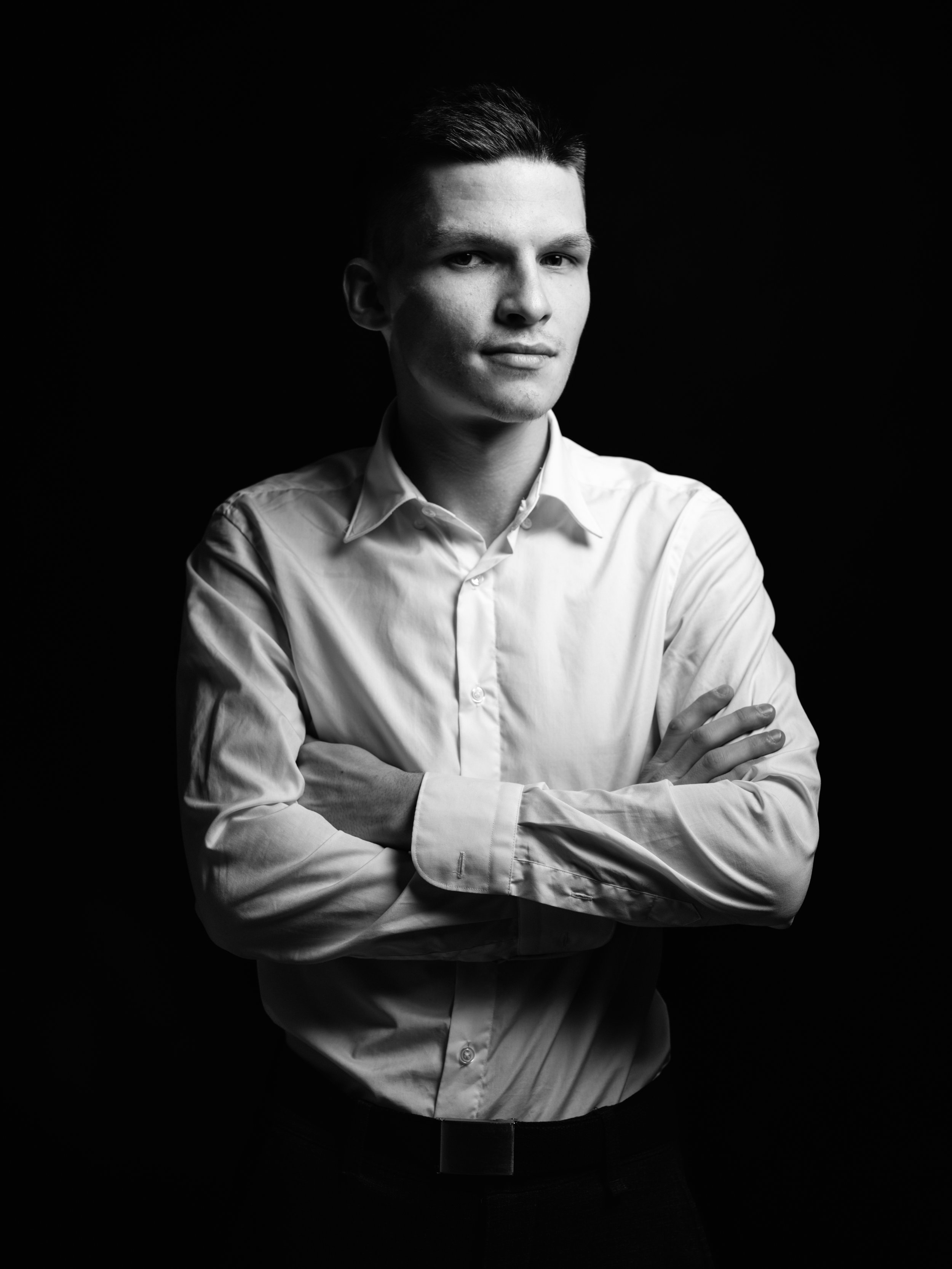 Black and white portrait of a young man with crossed arms, wearing a white dress shirt, looking confident against a dark background.