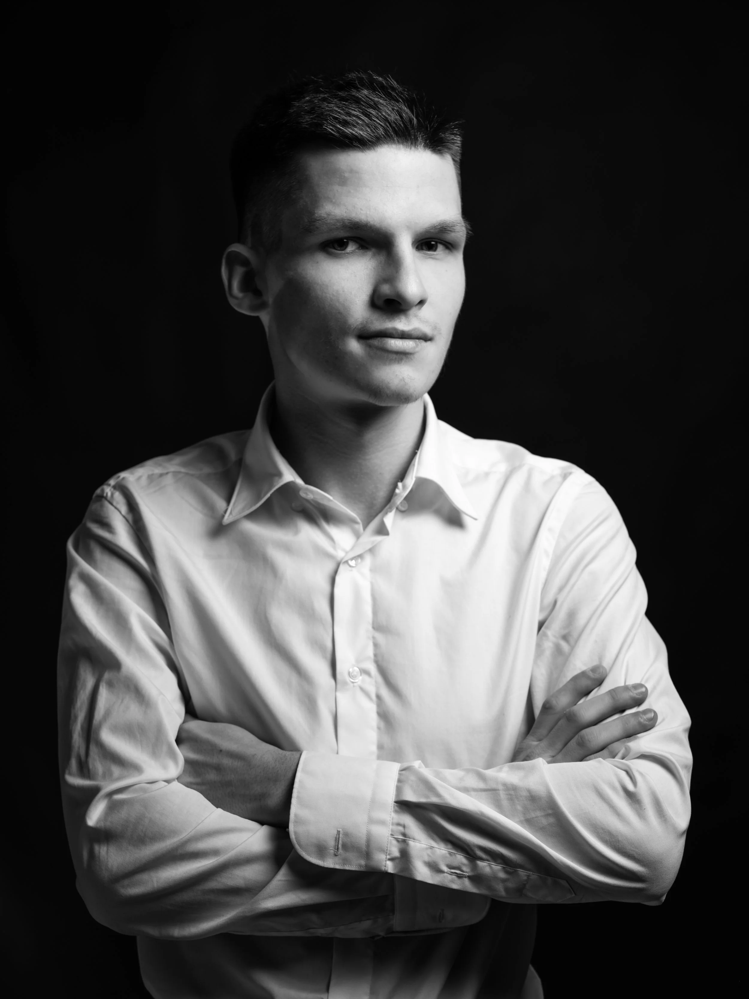 Black and white portrait of a young man with short dark hair, wearing a white dress shirt, crossed arms, against a dark background.
