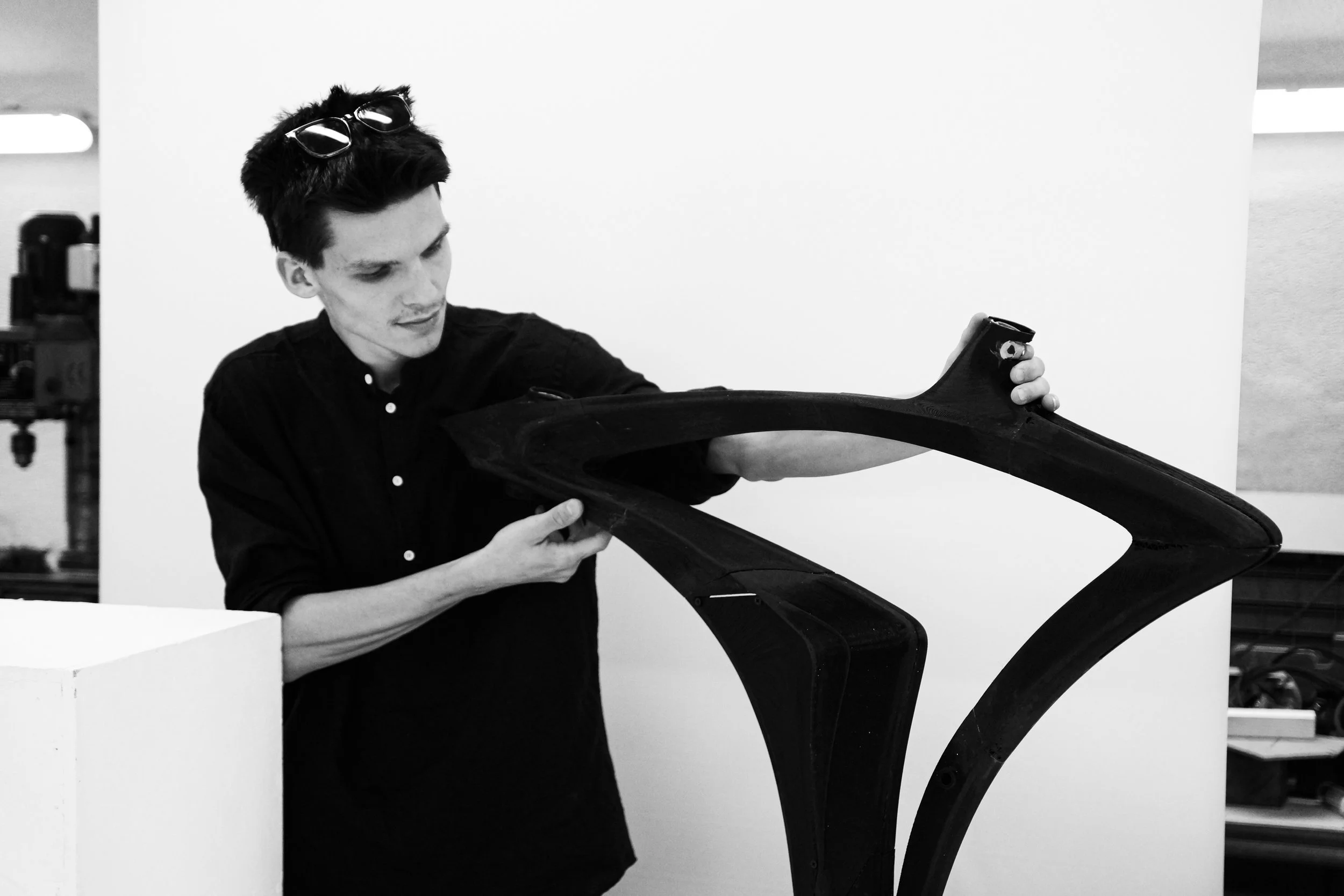 A young man examining a large black object, possibly a piece of equipment or part, in a workspace with a white wall background.