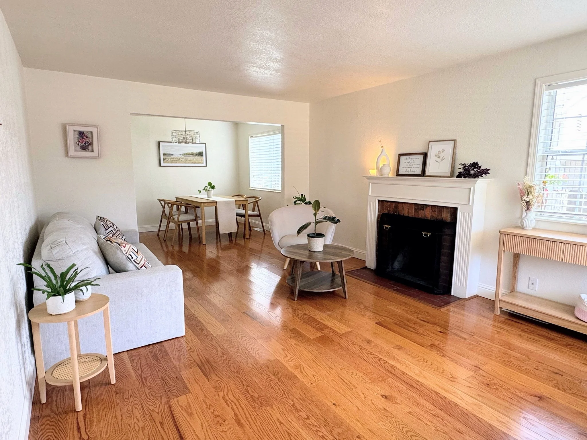 A living room and dining area with hardwood floors, a white sofa with patterned pillows, a small round side table with potted plants, a fireplace with decorative frames and vases on mantel, a wood dining table with chairs, and large windows with blinds.