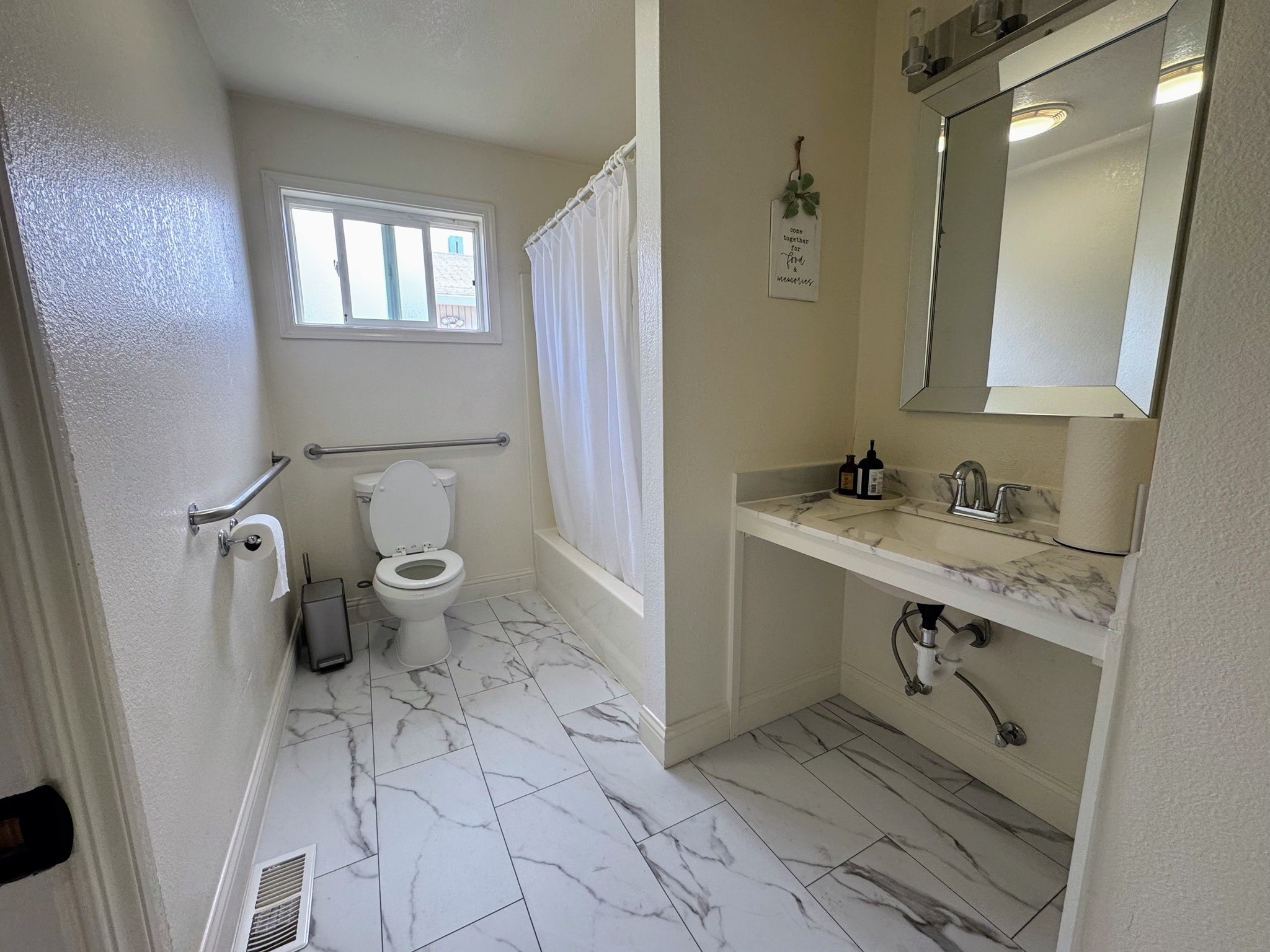 A small bathroom with white walls and marble tiled floor. Contains a toilet, a window, a shower with a white curtain, a white marble sink with a mirror above, and small bottles of soap or lotion on the sink.