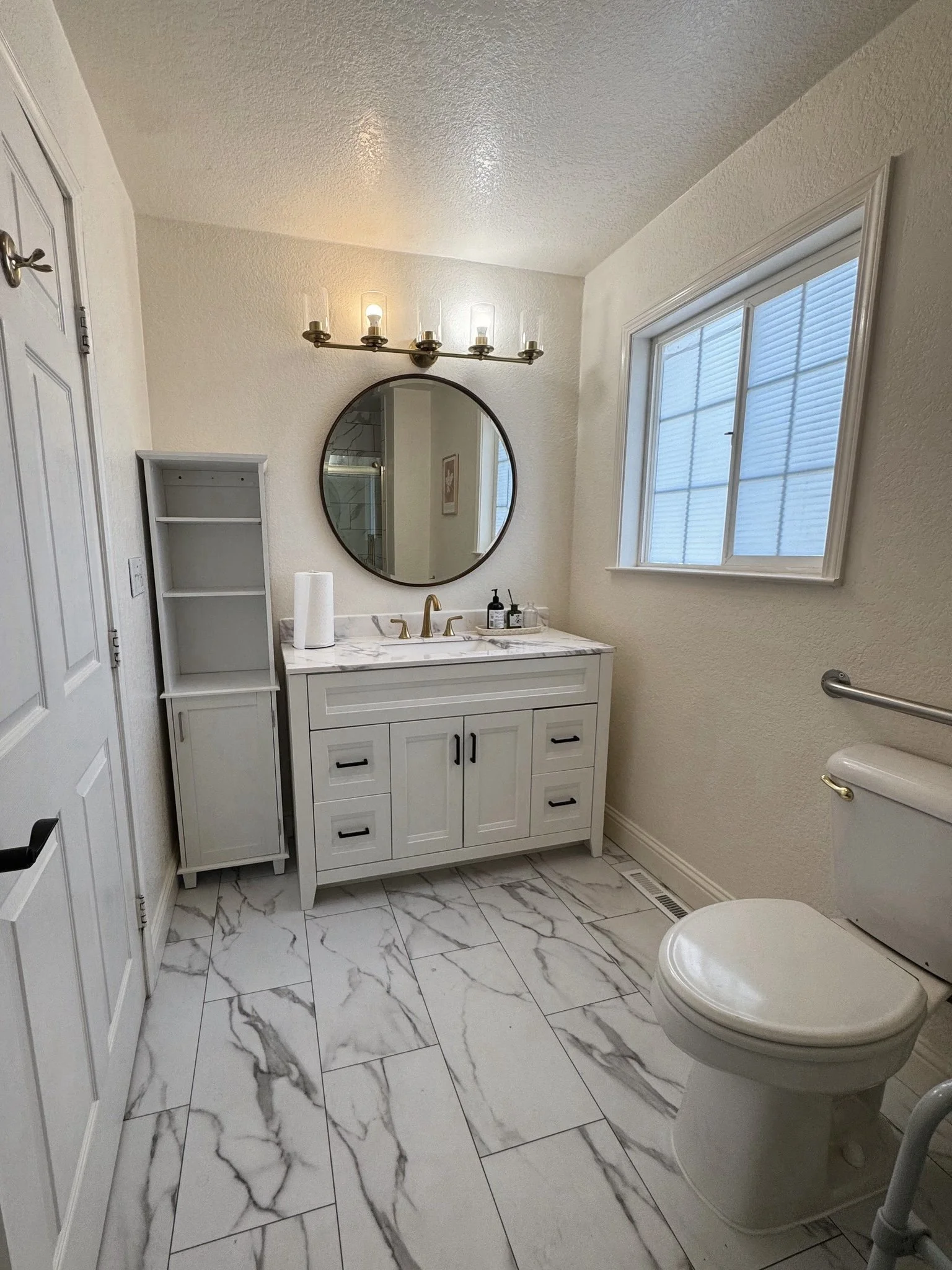 A bathroom with white walls and marble floor tiles. There is a white vanity with a marble countertop, a round mirror above it, and a black light fixture with multiple bulbs. To the right is a window with blinds, and to the left is a tall, open white 
