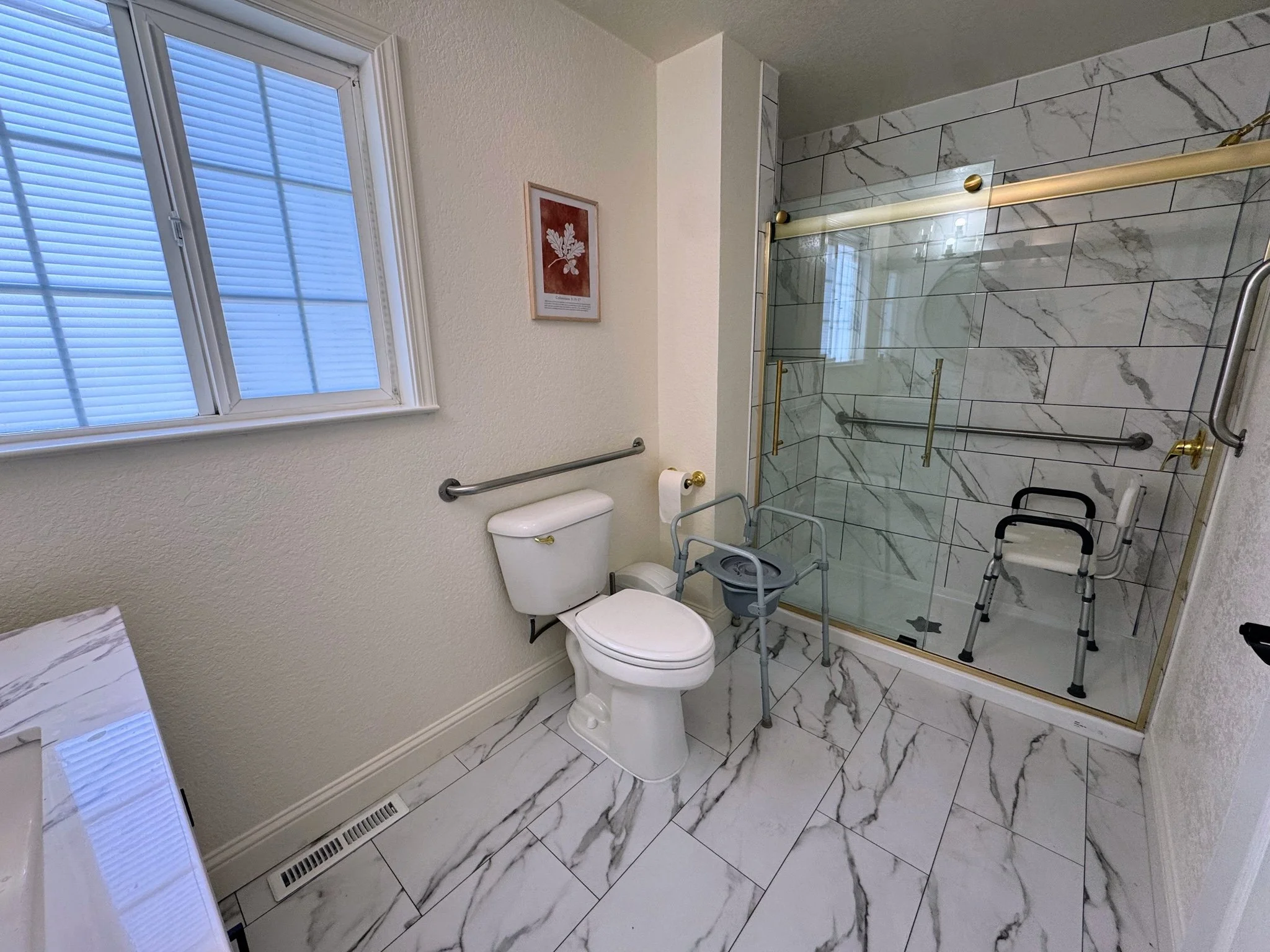 A bathroom with marble tile flooring and a marble tiled shower. There is a window with blinds, a framed artwork of a leaf, a white toilet, and two walkers.
